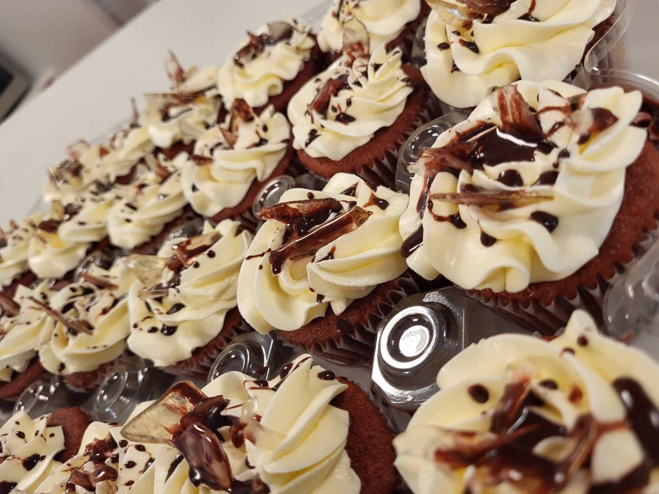 Multiple cupcakes with vanilla frosting, chocolate drizzle, and chocolate shavings on a white surface.