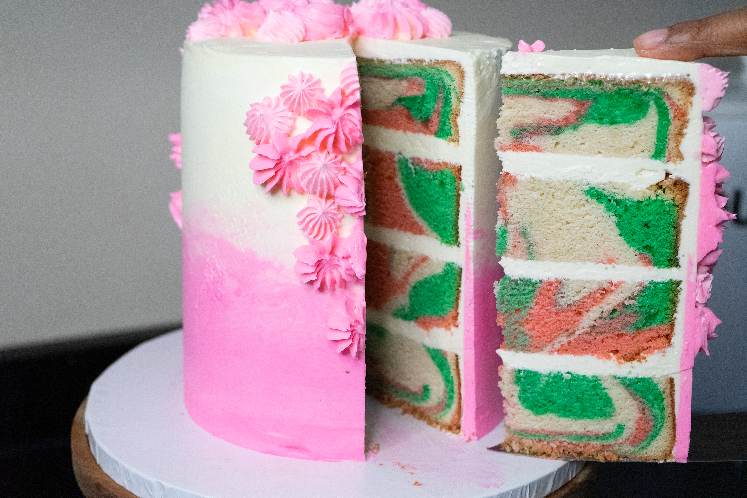 A partially sliced tiered cake with pastel pink ombre frosting and decorative pink buttercream flowers. The inside reveals a colorful swirl marble pattern of green, pink, and beige cake layers.