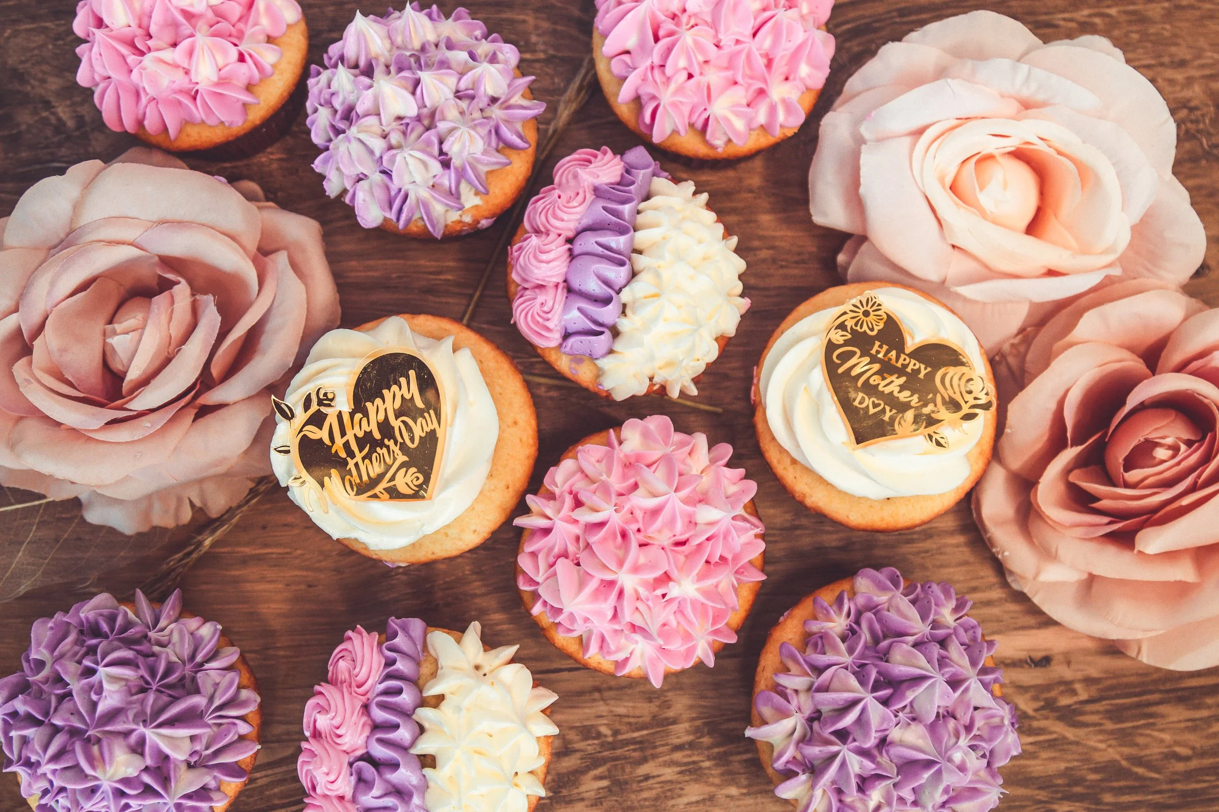 Decorative cupcakes with pink, purple, and white frosting arranged on a wooden surface, including some with 'Happy Mother's Day' edible toppers, and large pink and cream-colored rose-shaped frosting decorations.