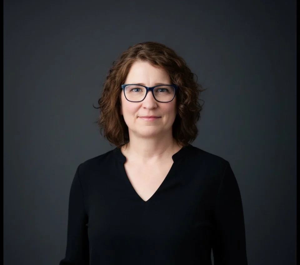 A woman with brown curly hair wearing glasses and a black shirt, looking at the camera against a dark gray background.