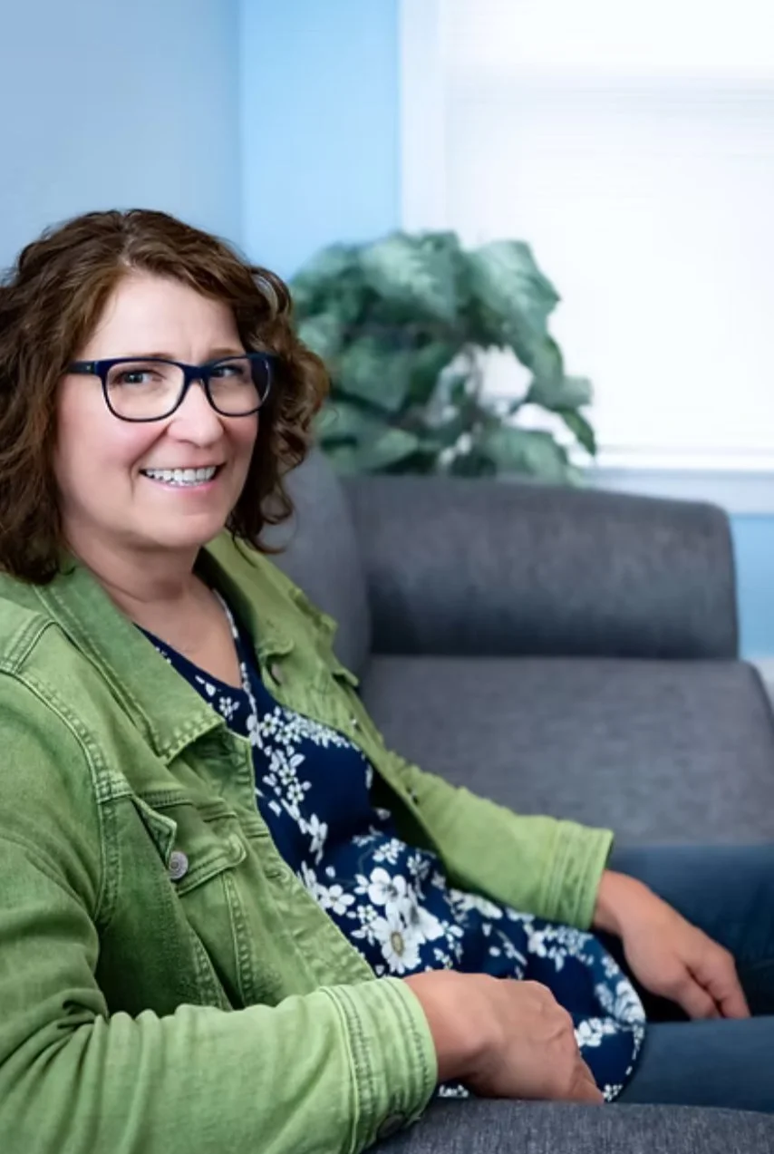 A woman with curly brown hair and glasses sitting on a gray couch in a bright room, smiling at the camera, wearing a green jacket and a navy blue dress with white floral pattern, with a large green houseplant and window in the background.
