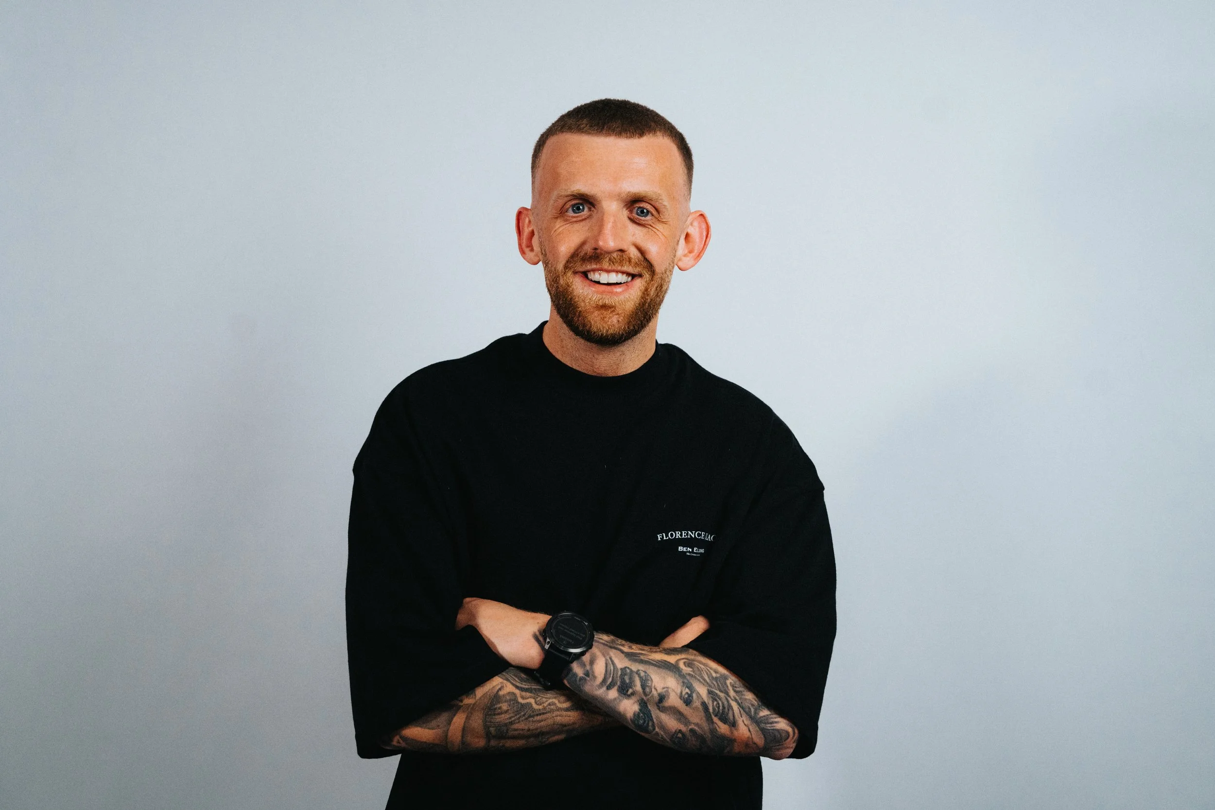 A man with short hair, a beard, and tattoos on his arm, wearing a black shirt and a watch, smiling and standing with arms crossed against a plain light background.