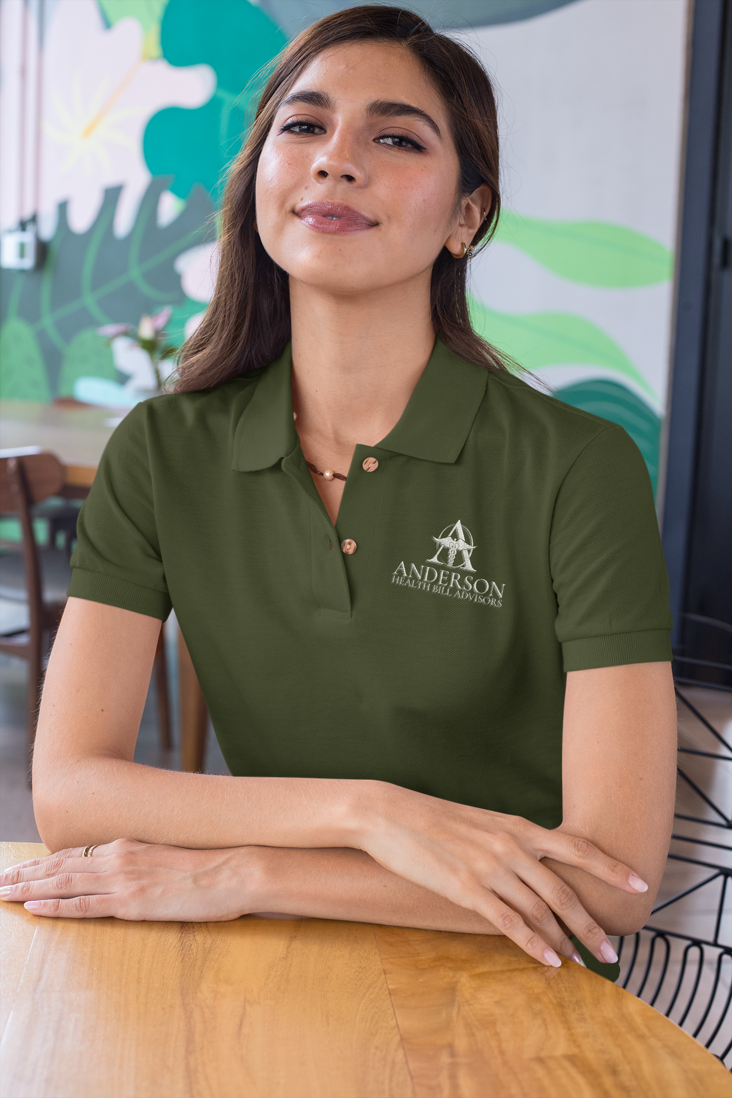A woman in a green polo shirt with 'Anderson Health Bill Advisors' logo, sitting at a wooden table in a casual setting.