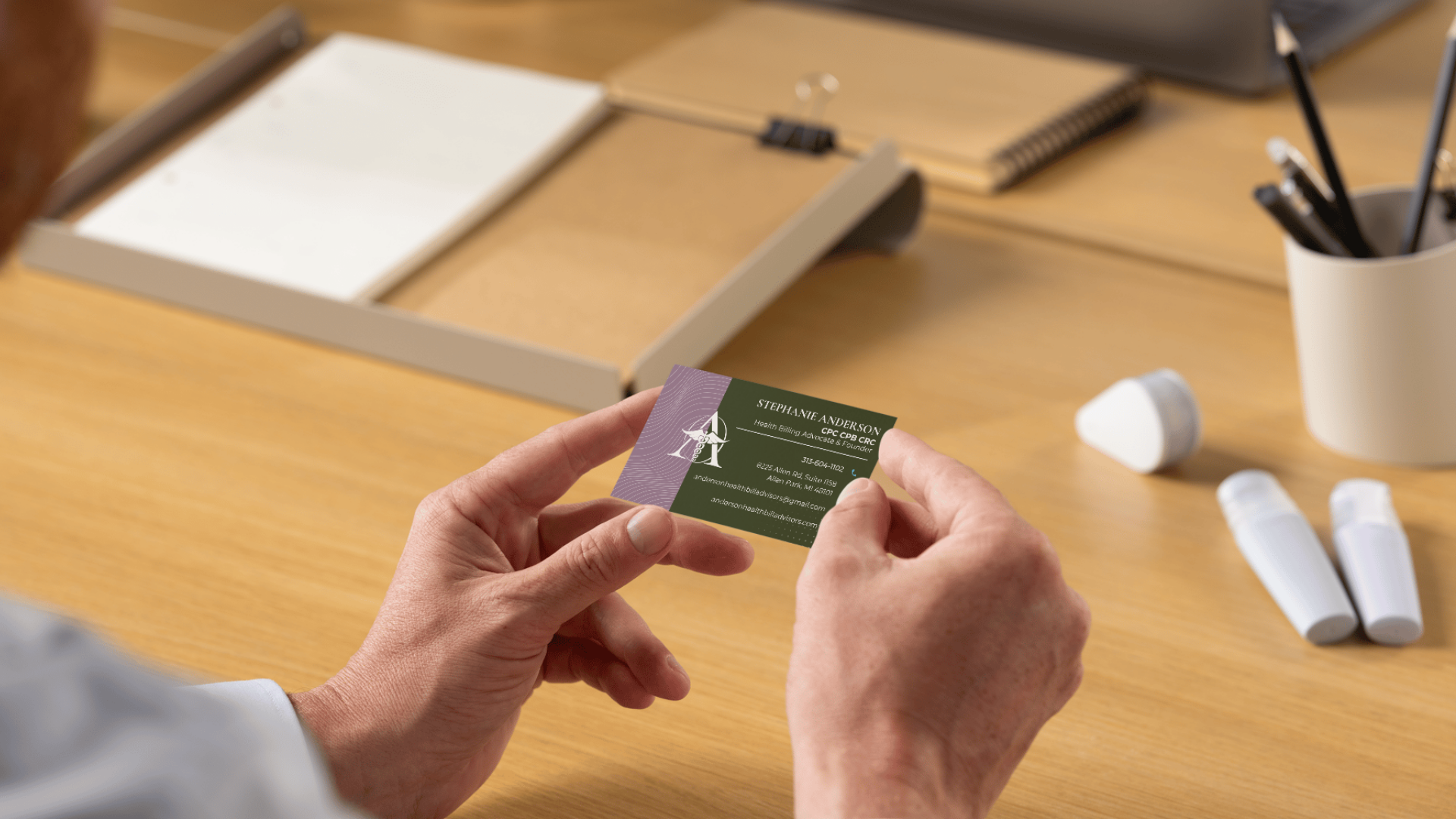 Person holding a business card with contact details at a desk with office supplies, including a paper cutter, two white bottles, and a cup filled with pens and pencils.