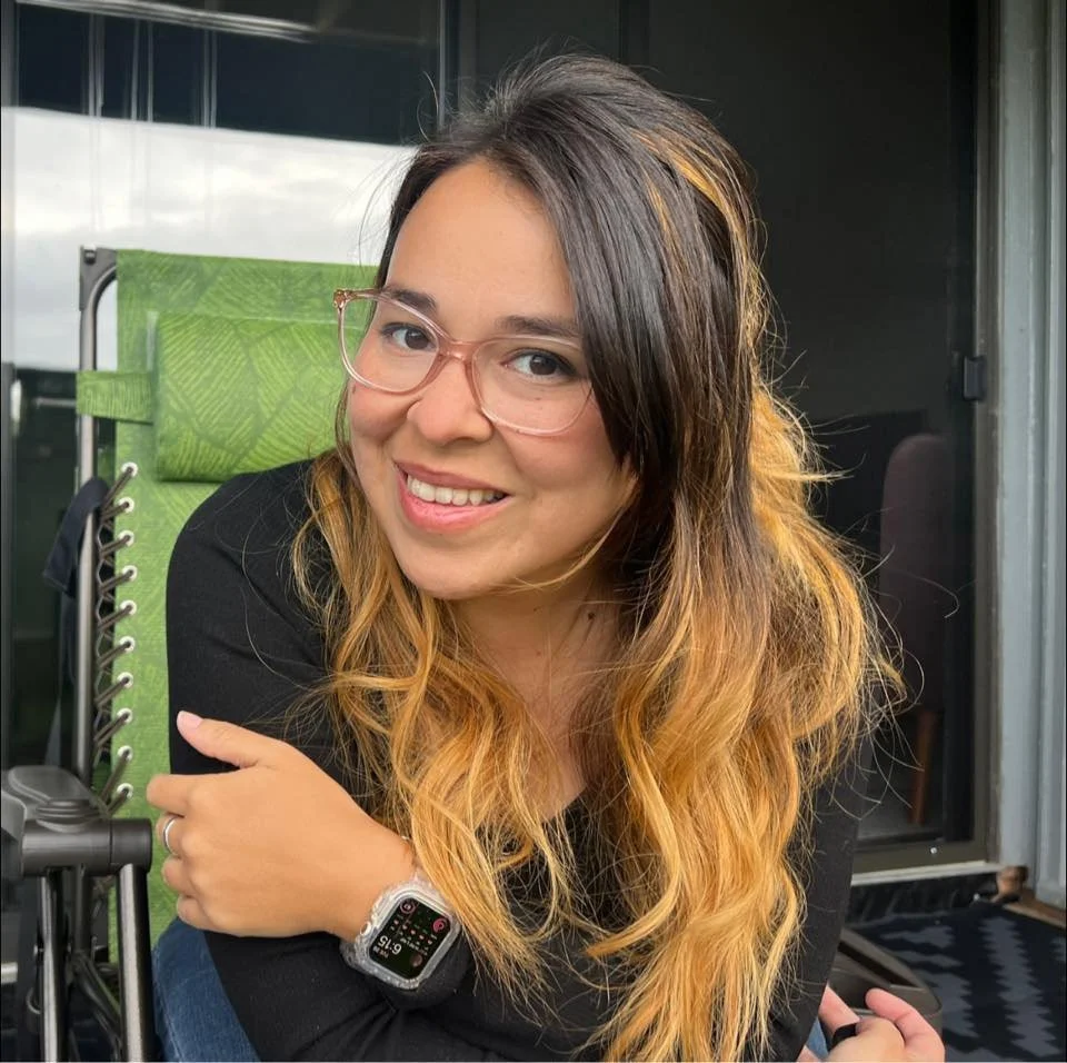 A young woman with glasses and long wavy hair smiling outdoors.