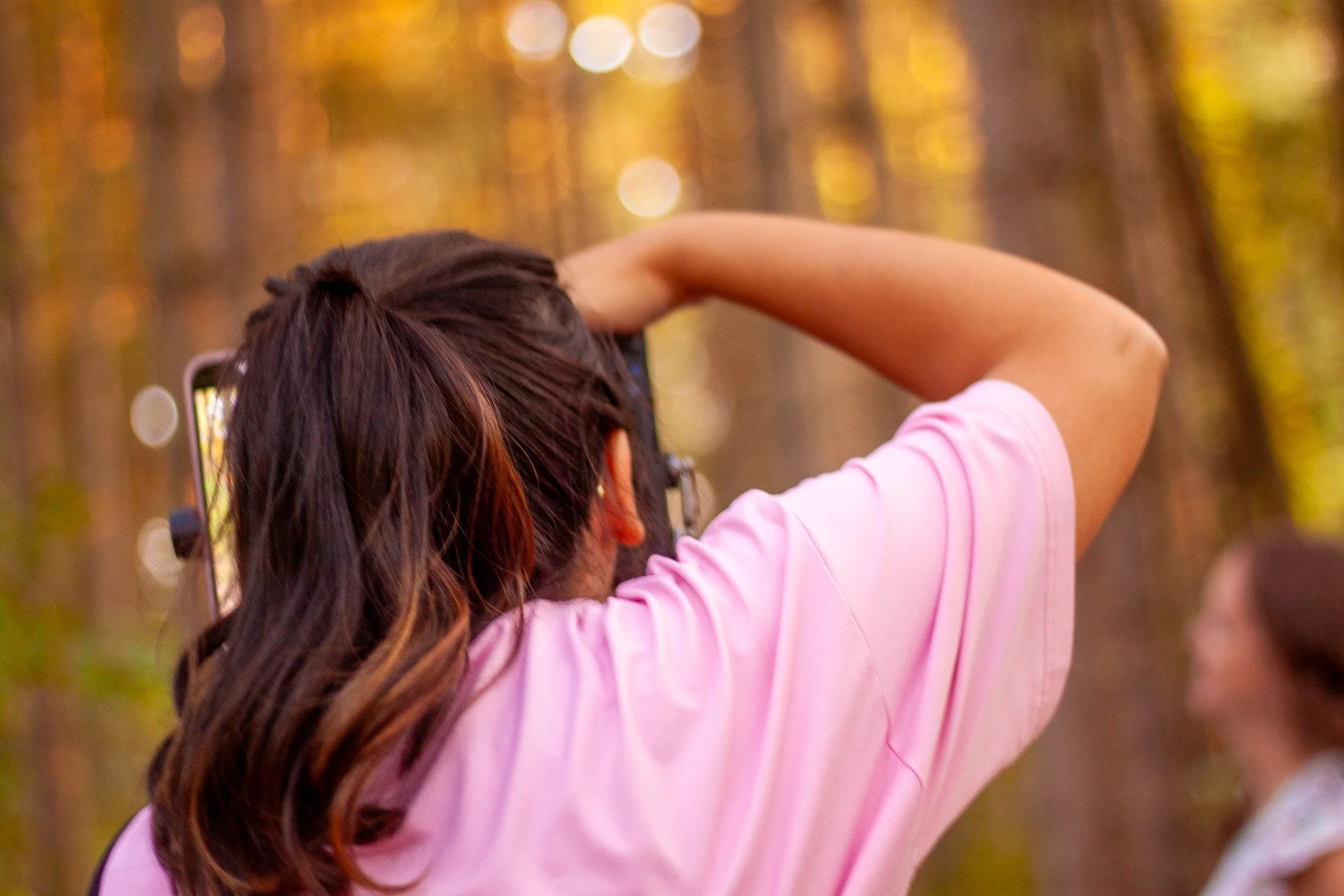 A person with long brown hair, wearing a pink shirt, takes a photo with a smartphone in a forest during autumn.