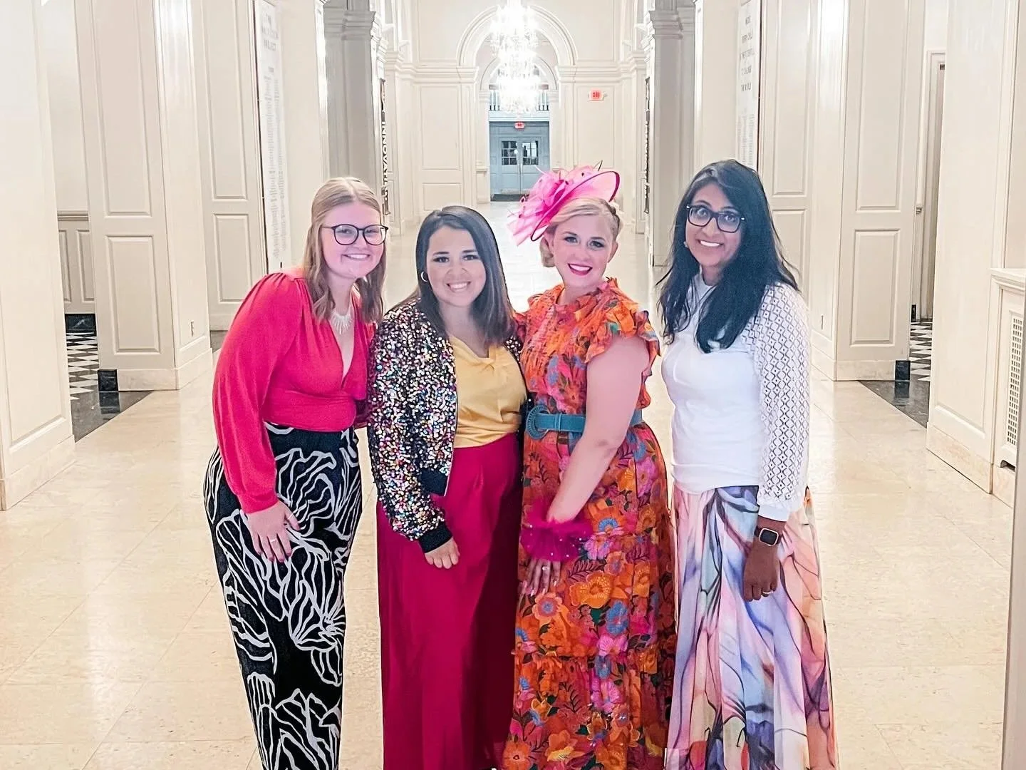 Four women standing together in a well-lit, elegant hallway, smiling at the camera. They are dressed in colorful, fashionable outfits, with one woman wearing a large pink hat and another with glasses.