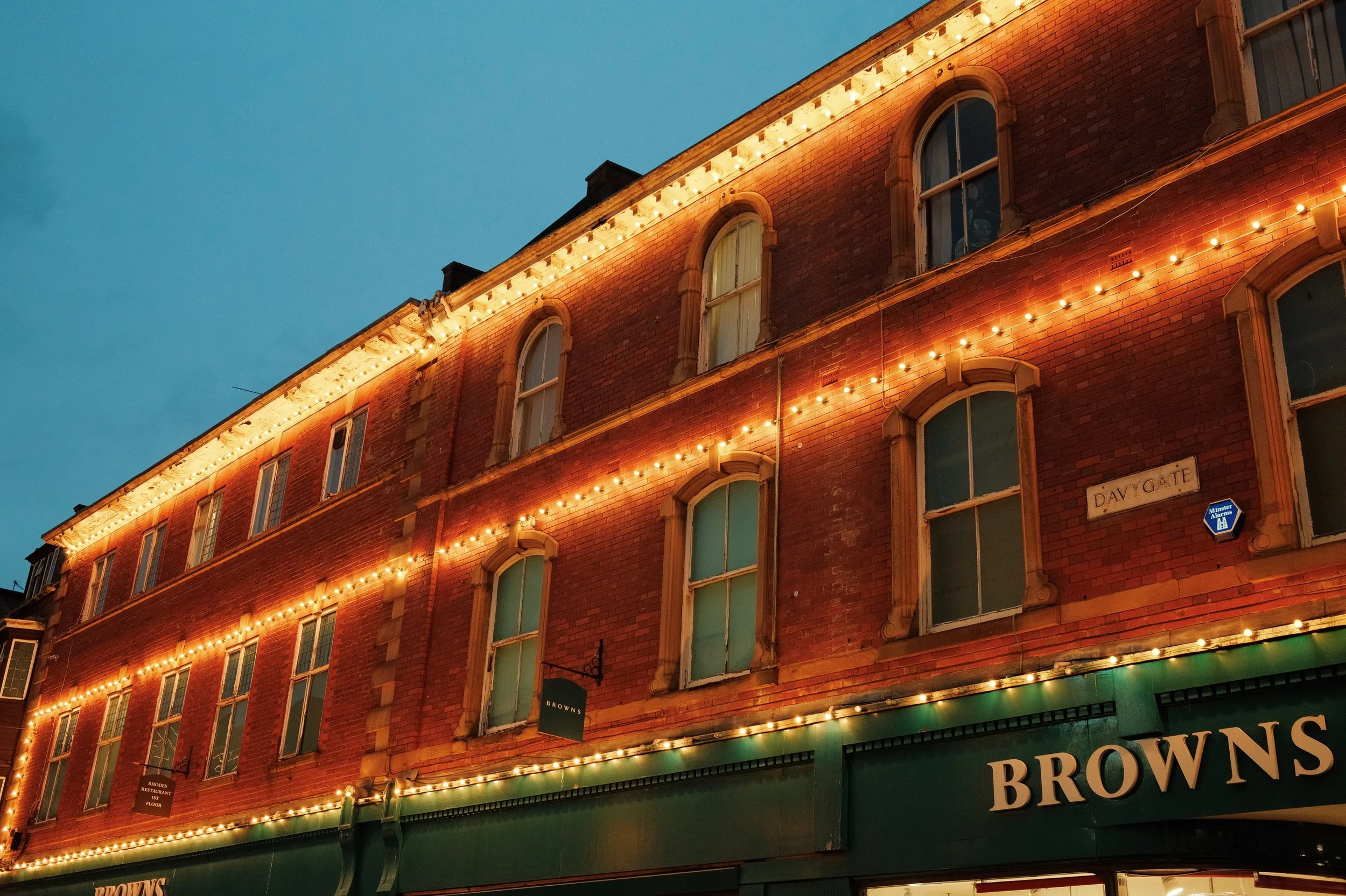 A multi-story brick building decorated with string lights along the roofline, at dusk. The building has arched windows and a green storefront with the name 'BROWNS'.