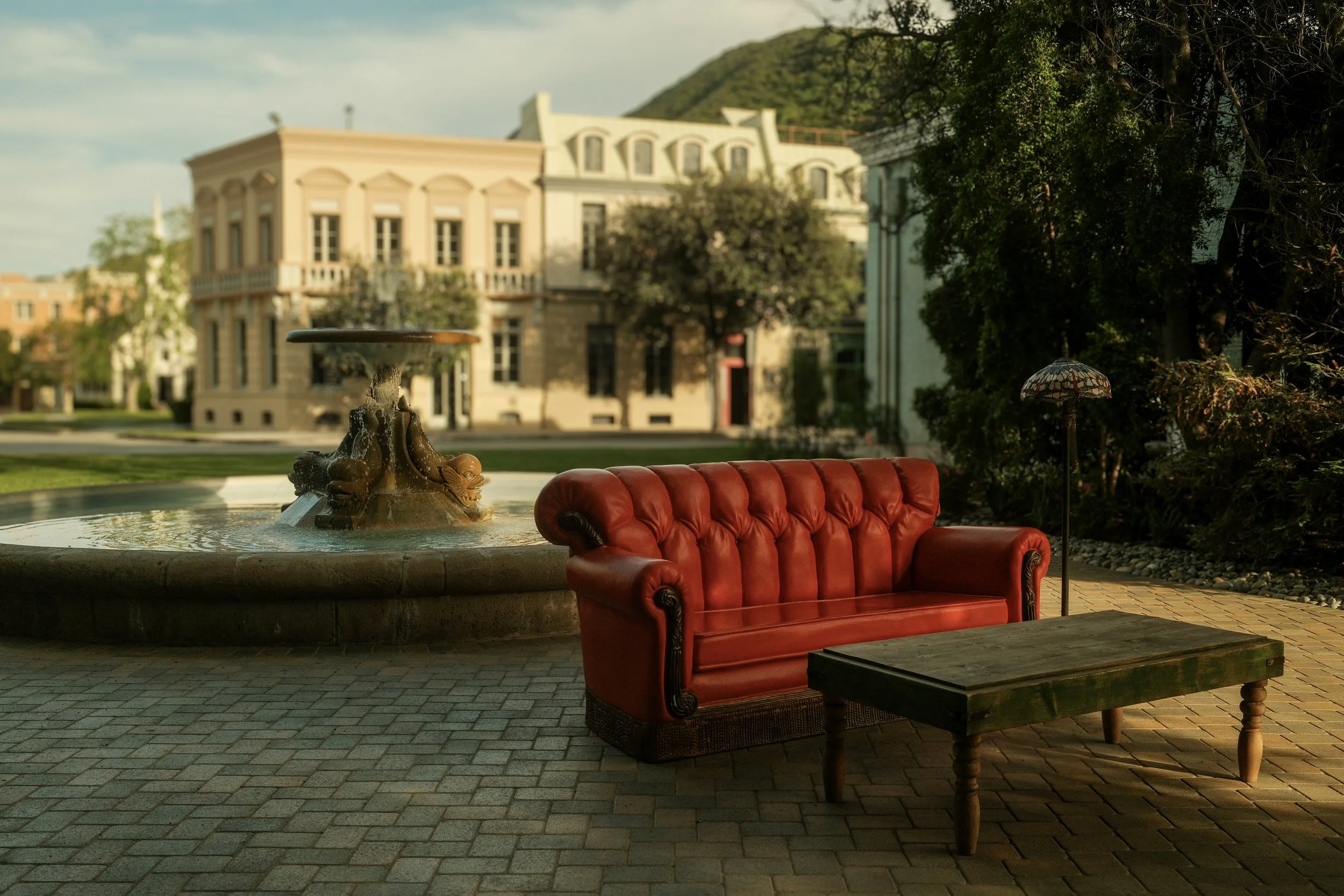 A vintage red leather sofa and a small wooden coffee table are outdoors near a fountain with a decorative sculpture. The background features historic buildings, trees, and a cloudy sky.