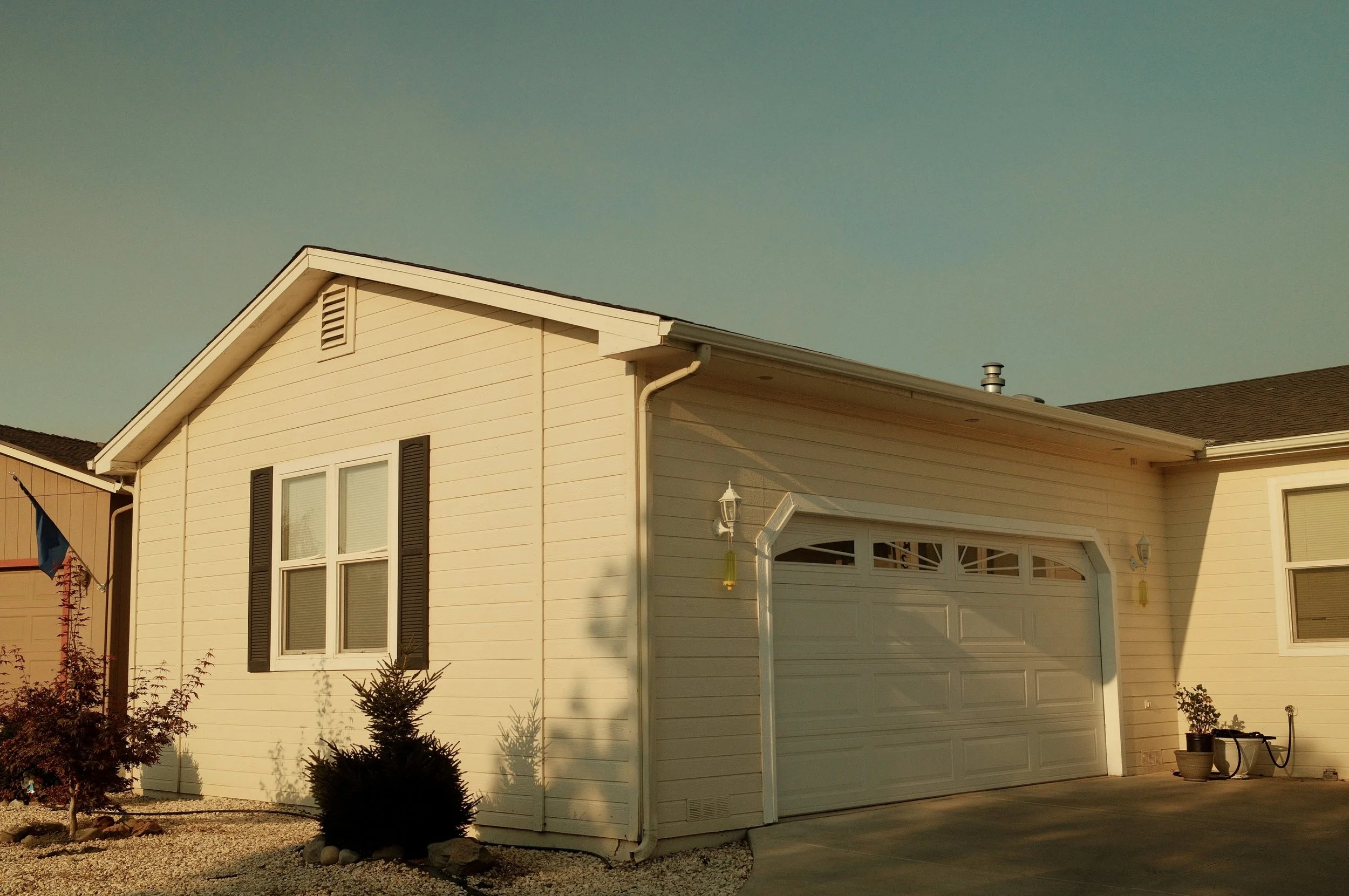 A yellow house with a white garage door, black shutters on the windows, and a small front yard with rocks and a tree.