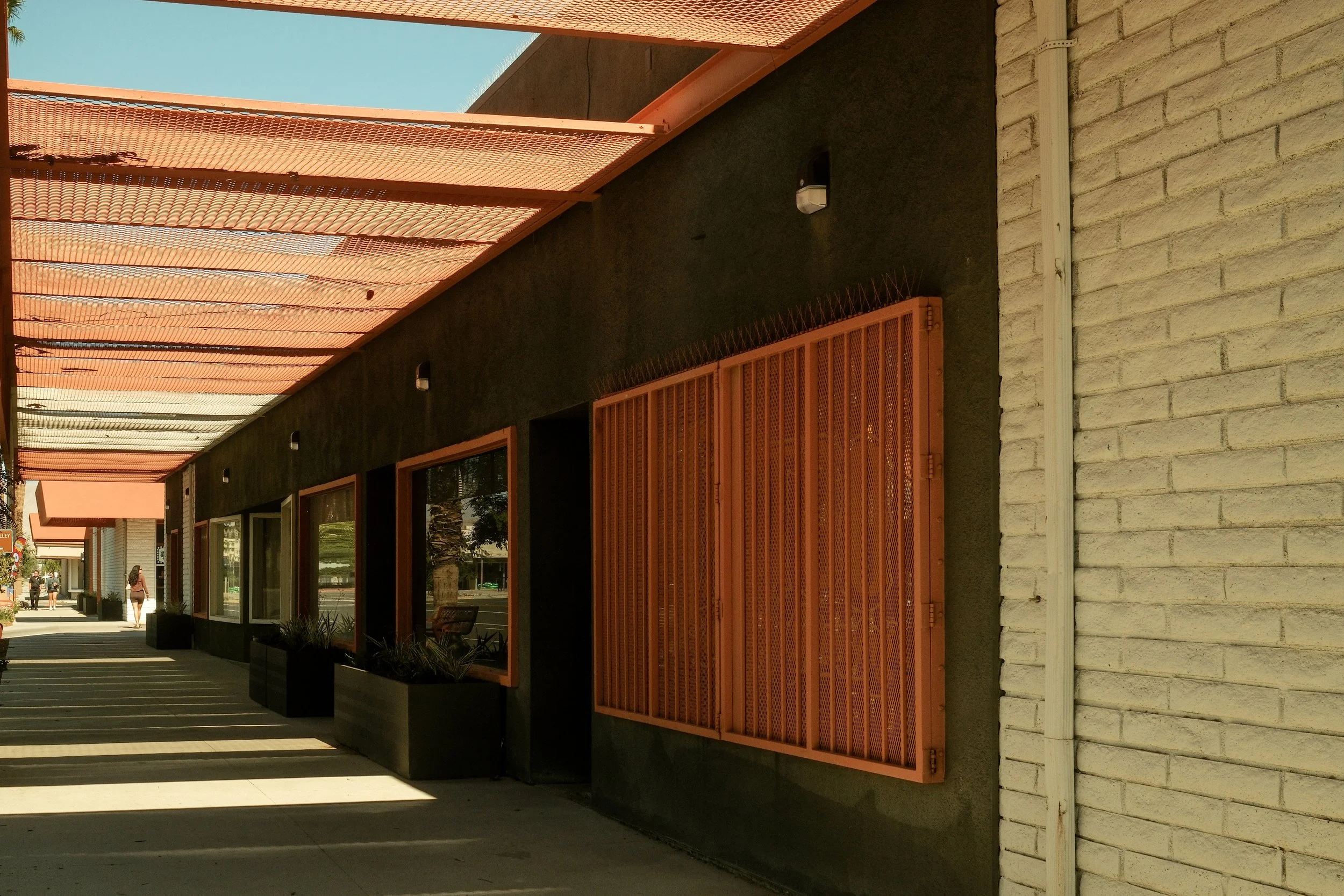 Sidewalk view of a modern commercial building with black walls, orange window frames, and orange metal awnings, with a person walking in the background.