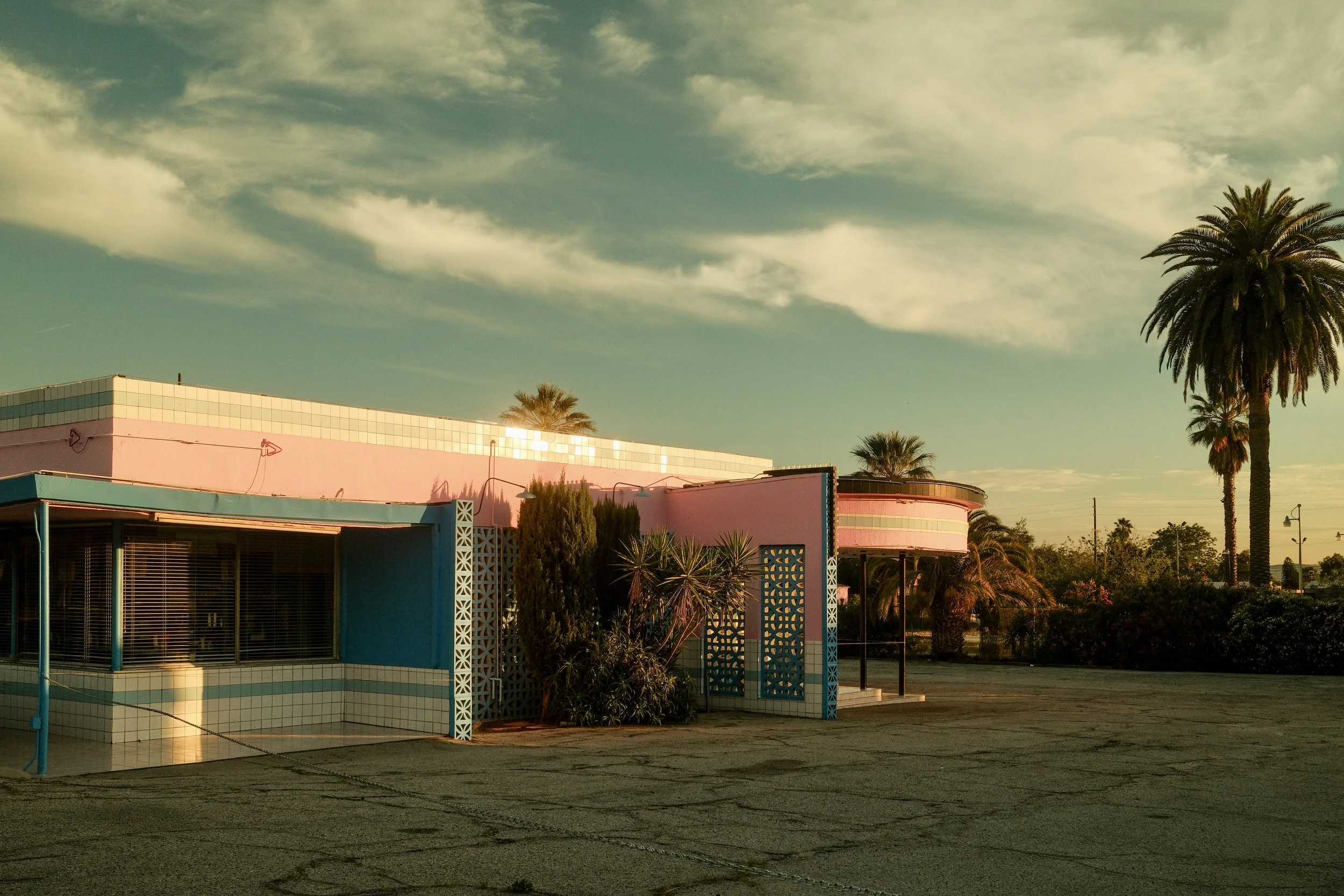 Retro-style building with pink and blue exterior, palm trees, and a partly cloudy sky at sunset.