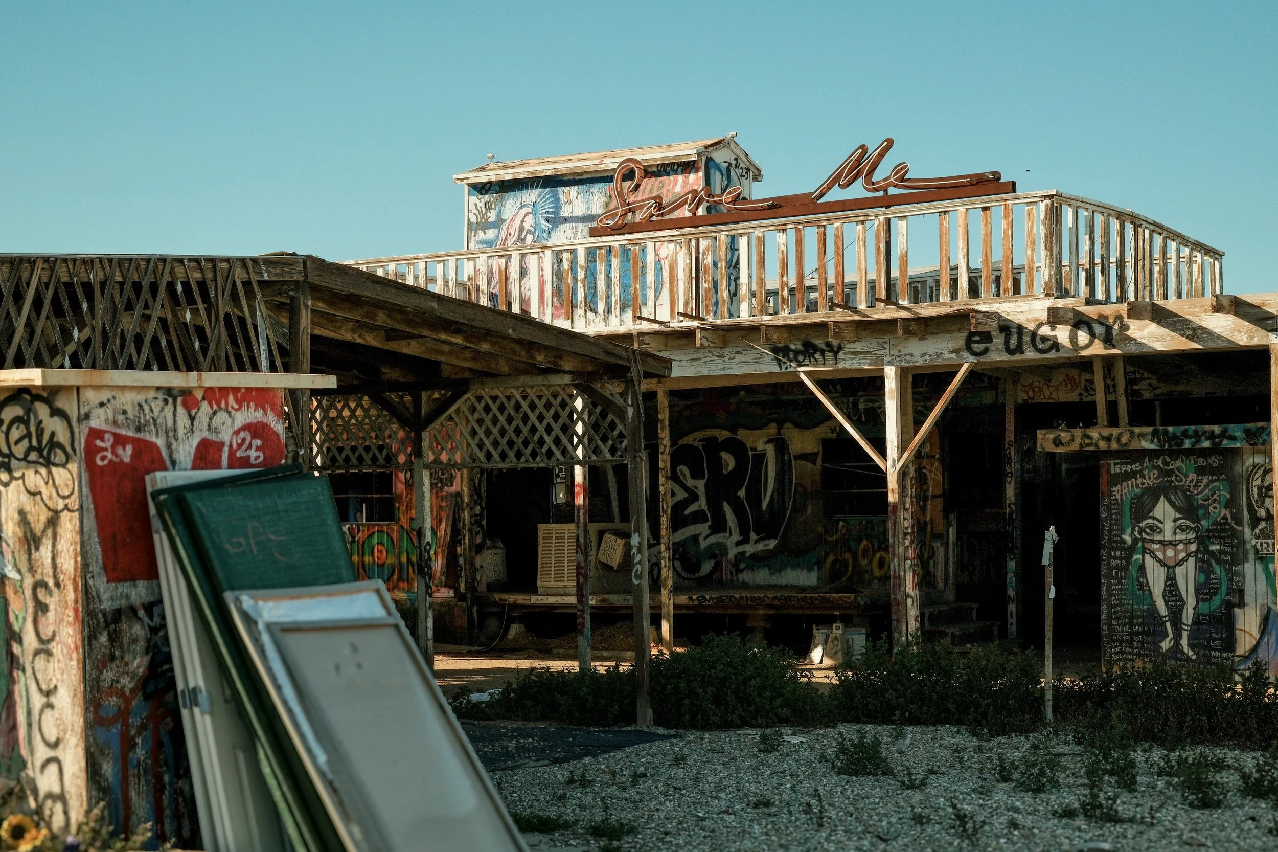 An old, graffiti-covered wooden structure with a sign that says 'Save Me' on a rooftop. The building appears abandoned, with peeling paint, graffiti art, and a clear blue sky in the background.