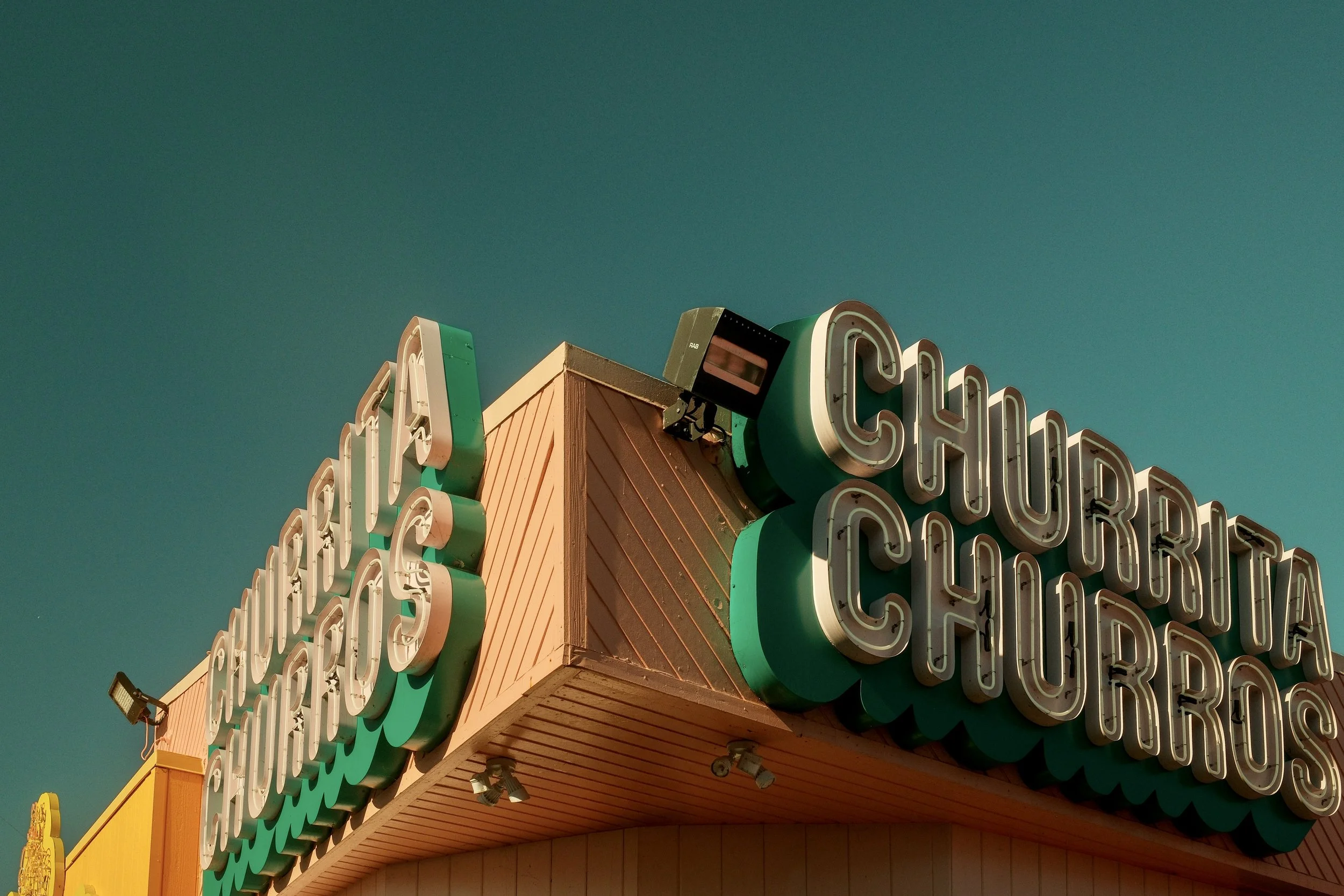 Close-up of a vintage sign with the words 'Churros' and 'Chocolate' in large, illuminated letters on a building exterior against a clear blue sky.