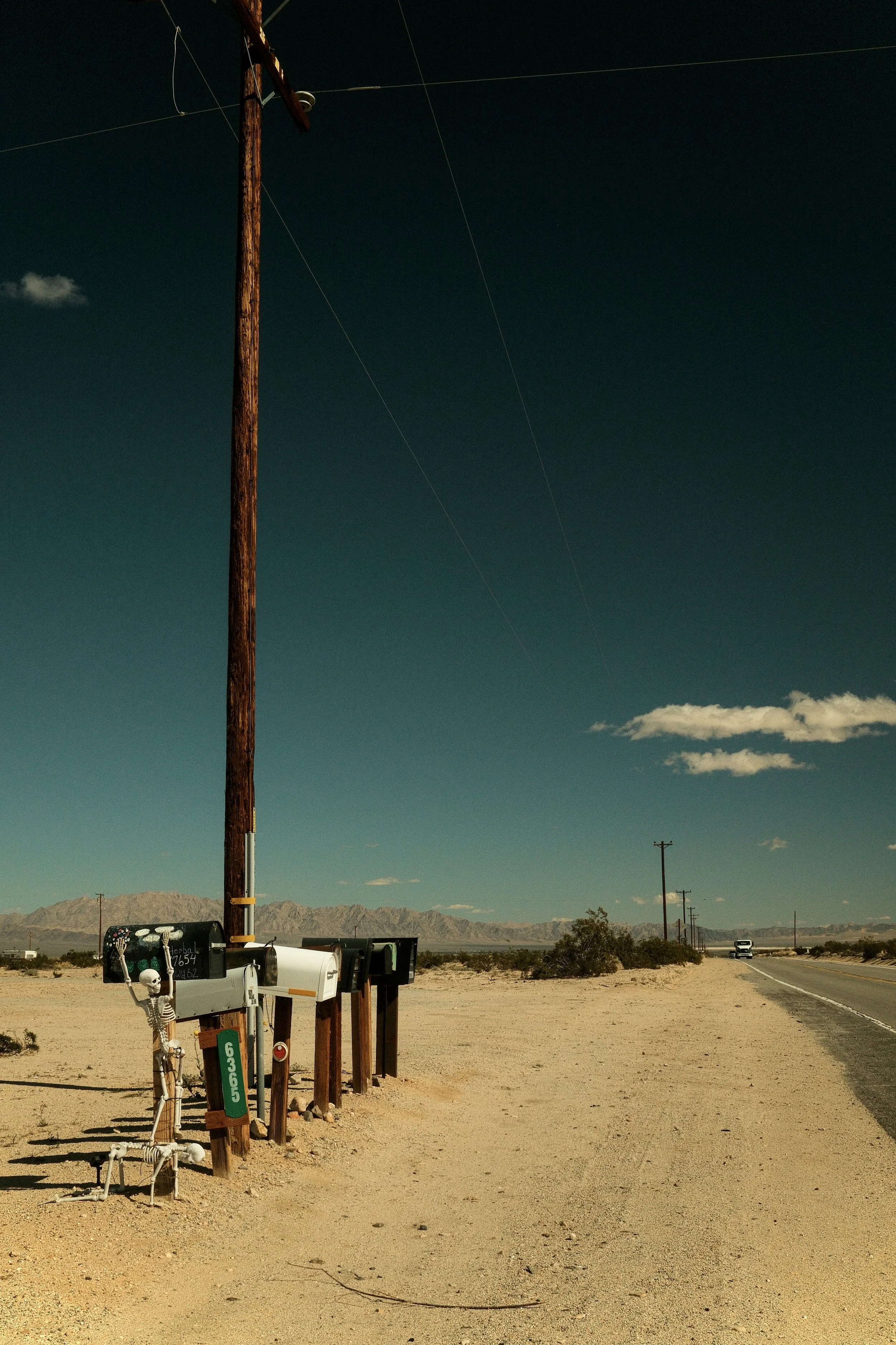 Desert highway scene with a wooden utility pole, multiple mailboxes, and a skeleton decoration on one mailbox. Mountains in the distance and a car approaching on the curved road.