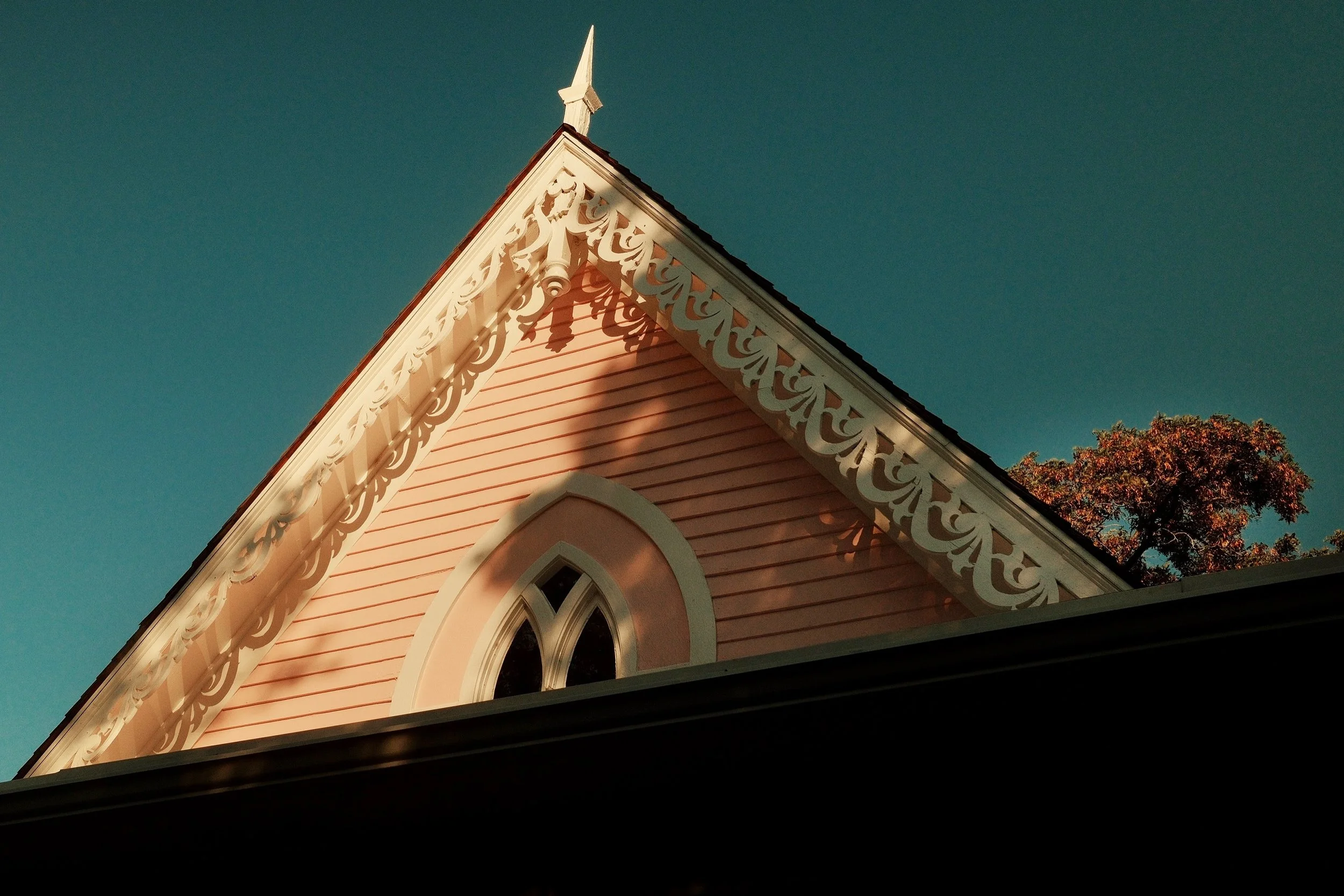 The corner of a pink Victorian-style house with white decorative trim, a pointed peak, and a Gothic-style window, with a tree and sky in the background.