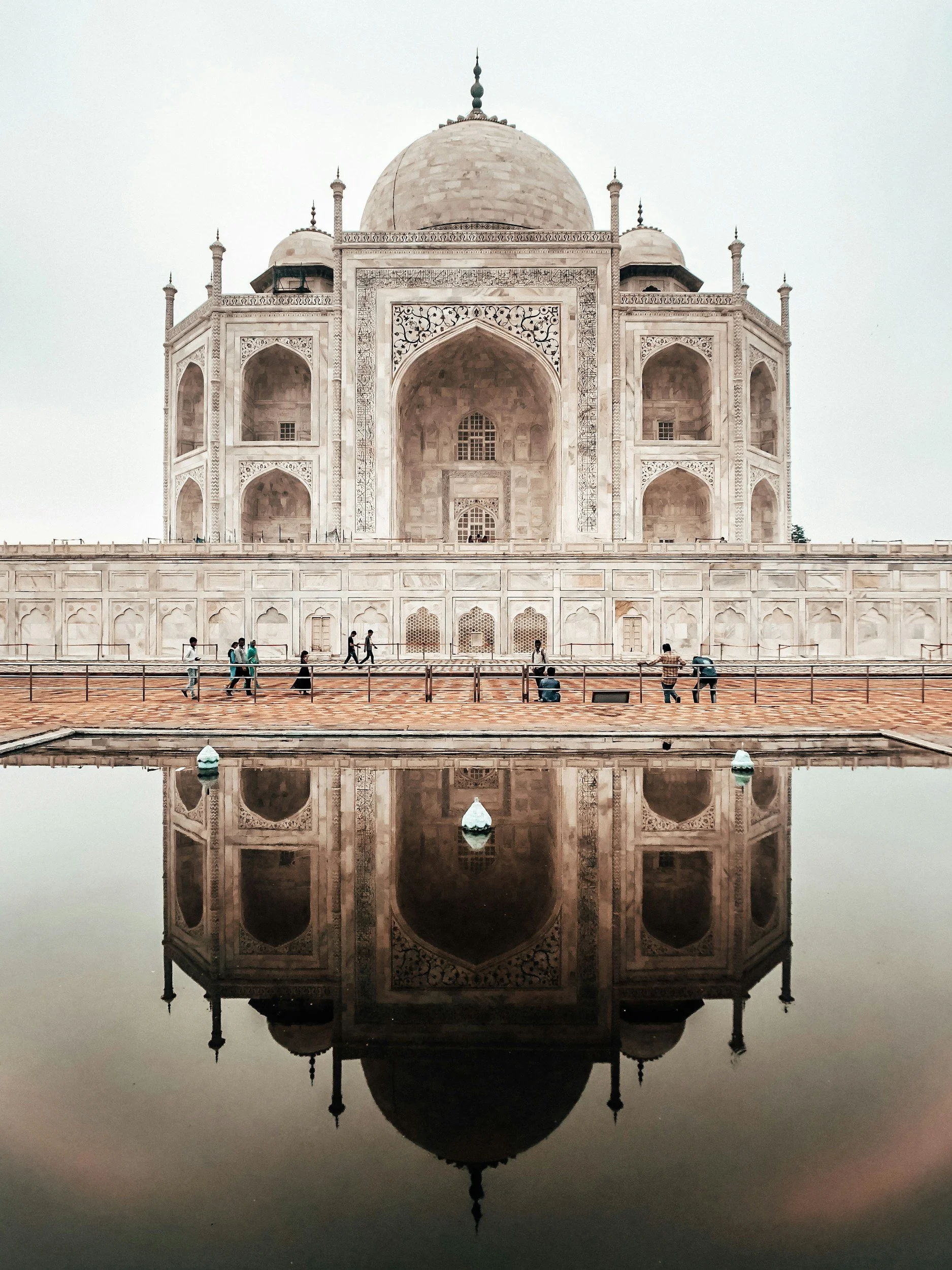 The Taj Mahal, a white marble mausoleum in India, with its reflection visible in a water pond in front of it, and visitors walking along the pathway.