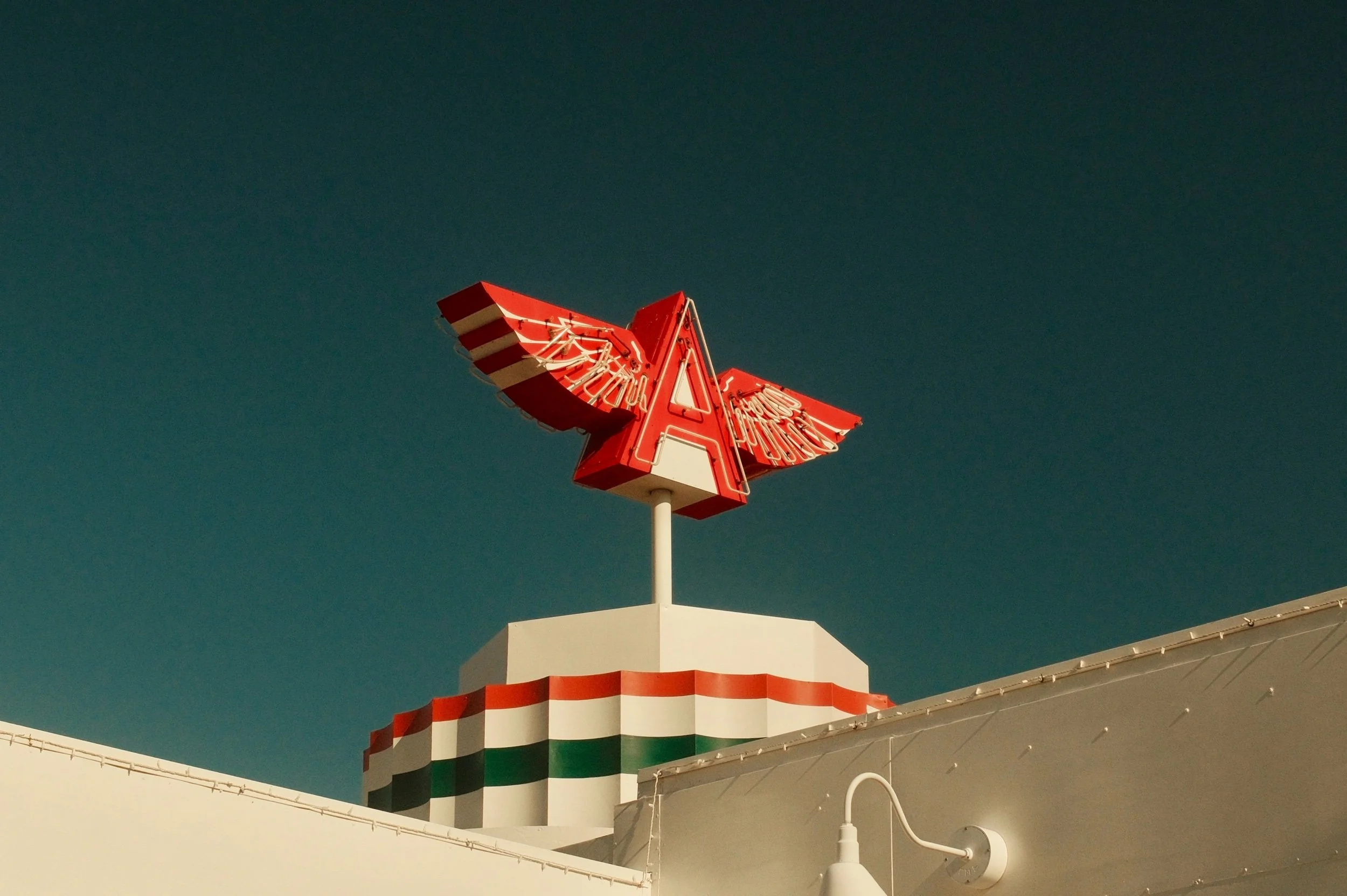 A red and white neon sign with wings, mounted on a pole, on top of a building decorated with a red, white, and green stripe pattern, against a dark sky.