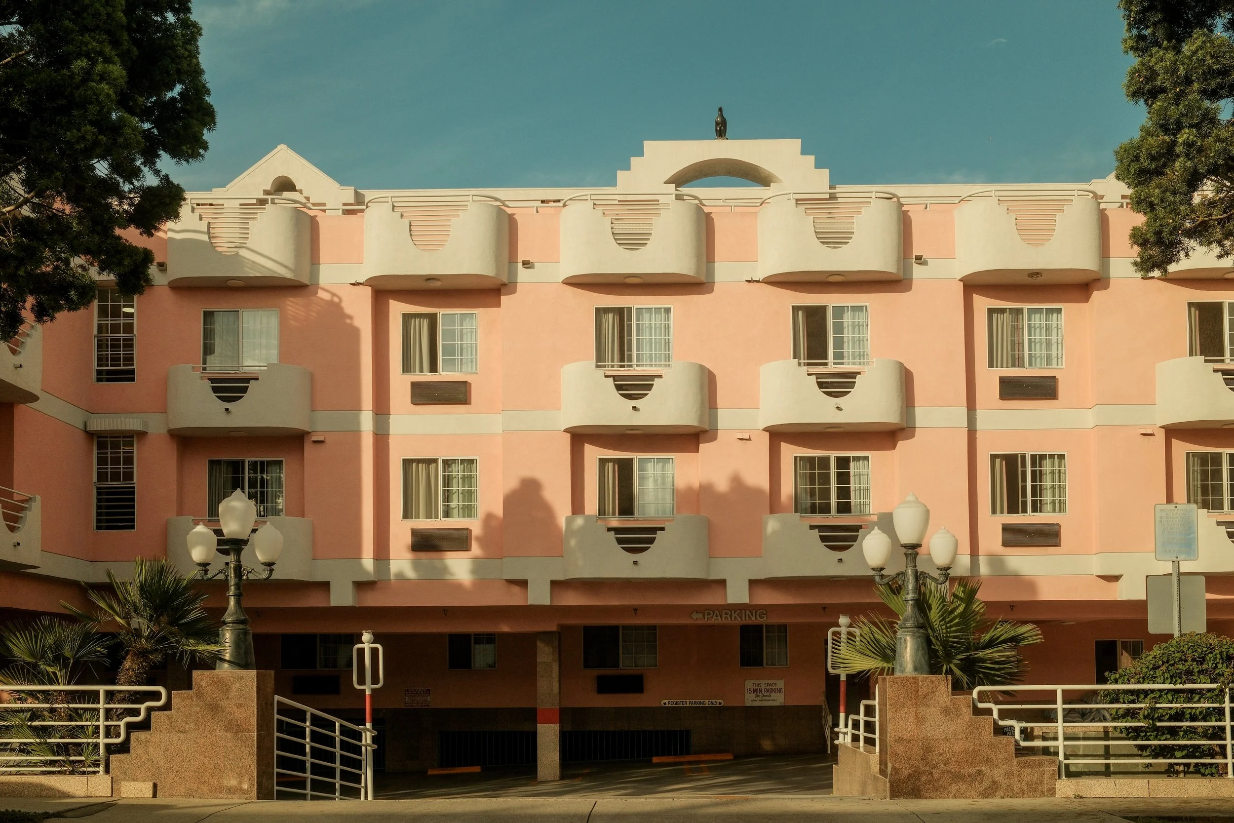 Pink multi-story motel building with small balconies and decorative architectural features, parking area at the ground level, trees and street lamps in front, clear blue sky in the background.