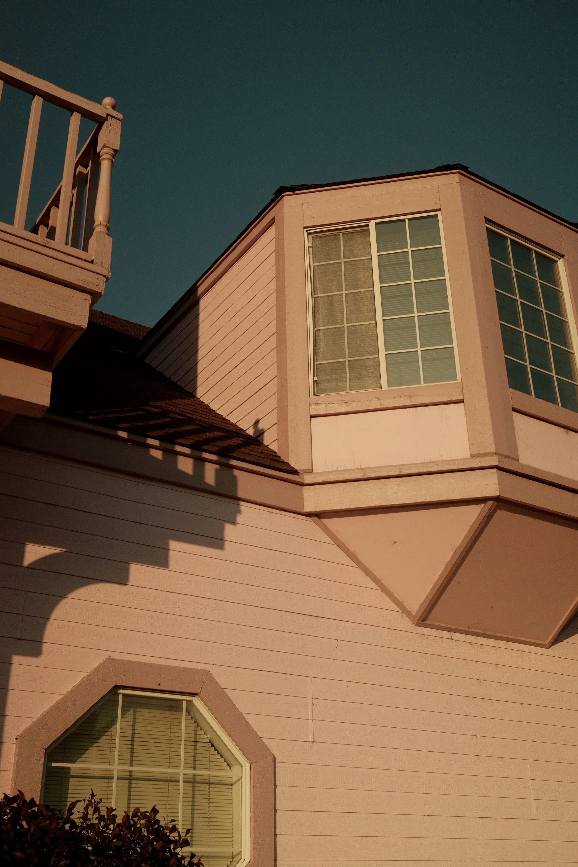 Close-up of a home with pink siding, large bay windows, and a balcony with a wooden railing under a clear sky.