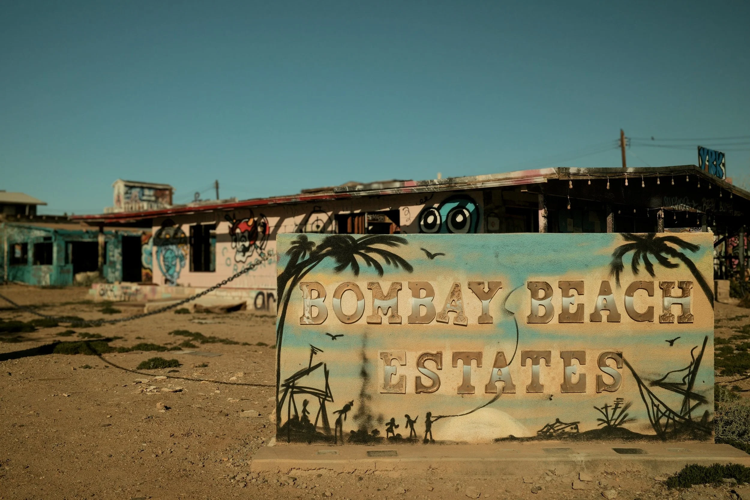 Sign that says 'Bombay Beach Estates' with painted palm trees, birds, and silhouettes of people, central to a graffiti-covered building in the background.