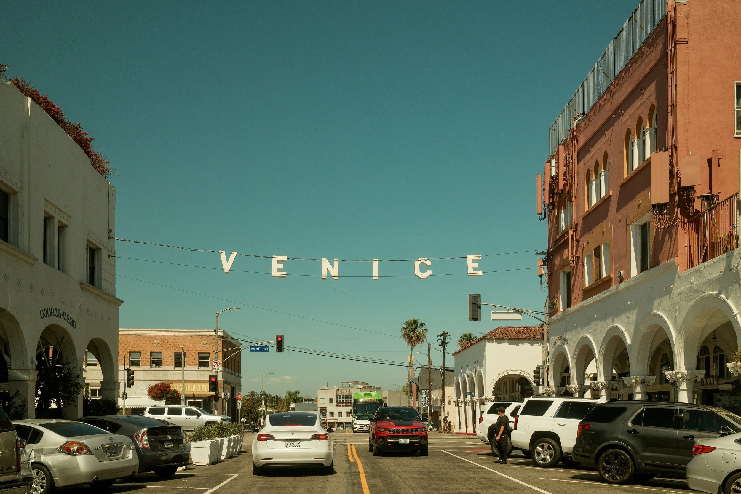 Street scene in Venice with parked cars, palm trees, and colorful buildings, and a string of letters spelling out 'VENICE' overhead.