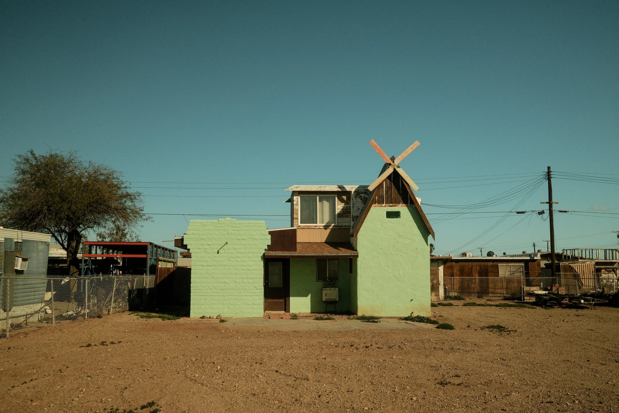 A small pastel green house with a windmill on top, situated on an arid lot with a clear blue sky in the background.
