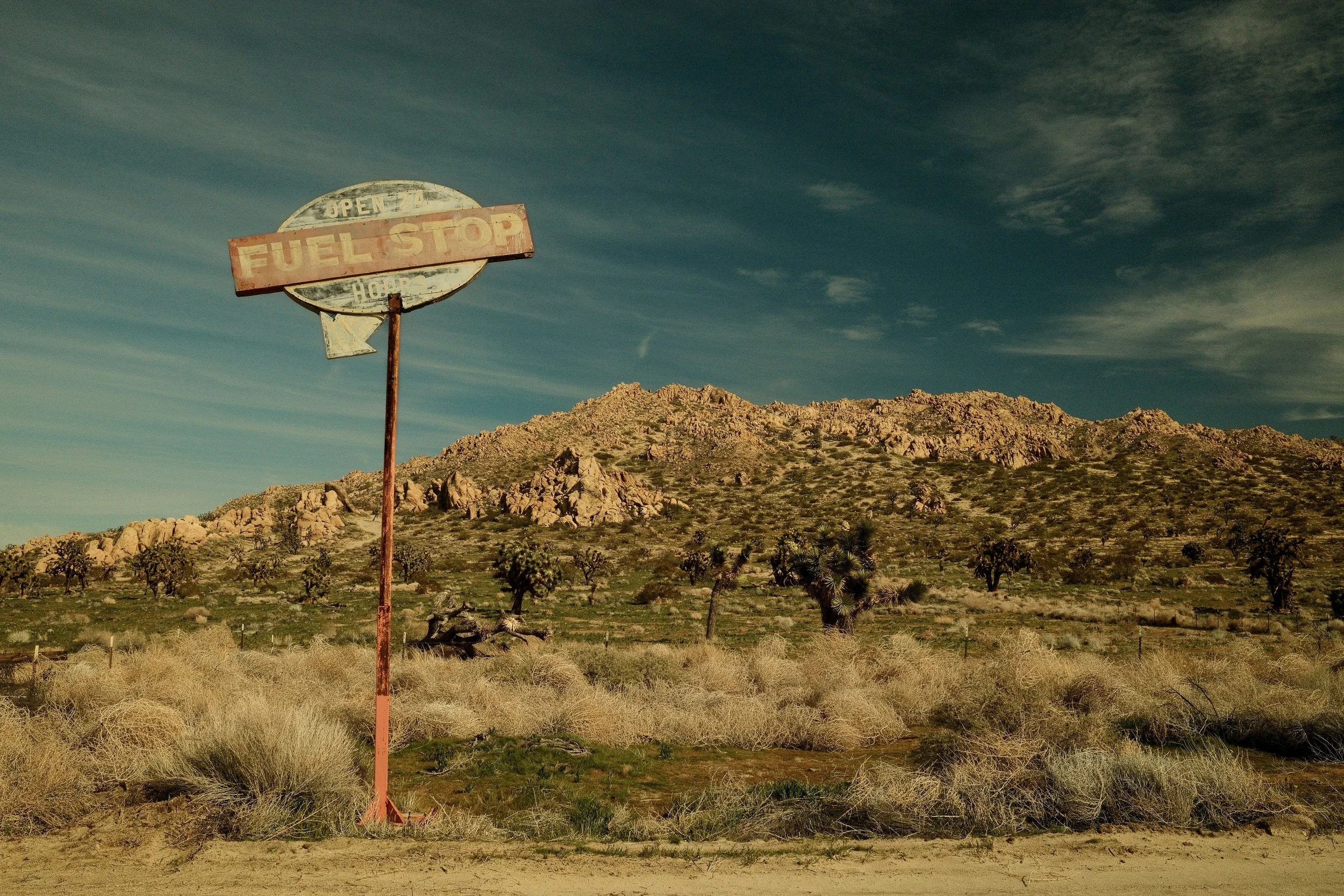 Old, rusty sign in a desert landscape with mountains and Joshua trees, reading 'Fuel Stop'.