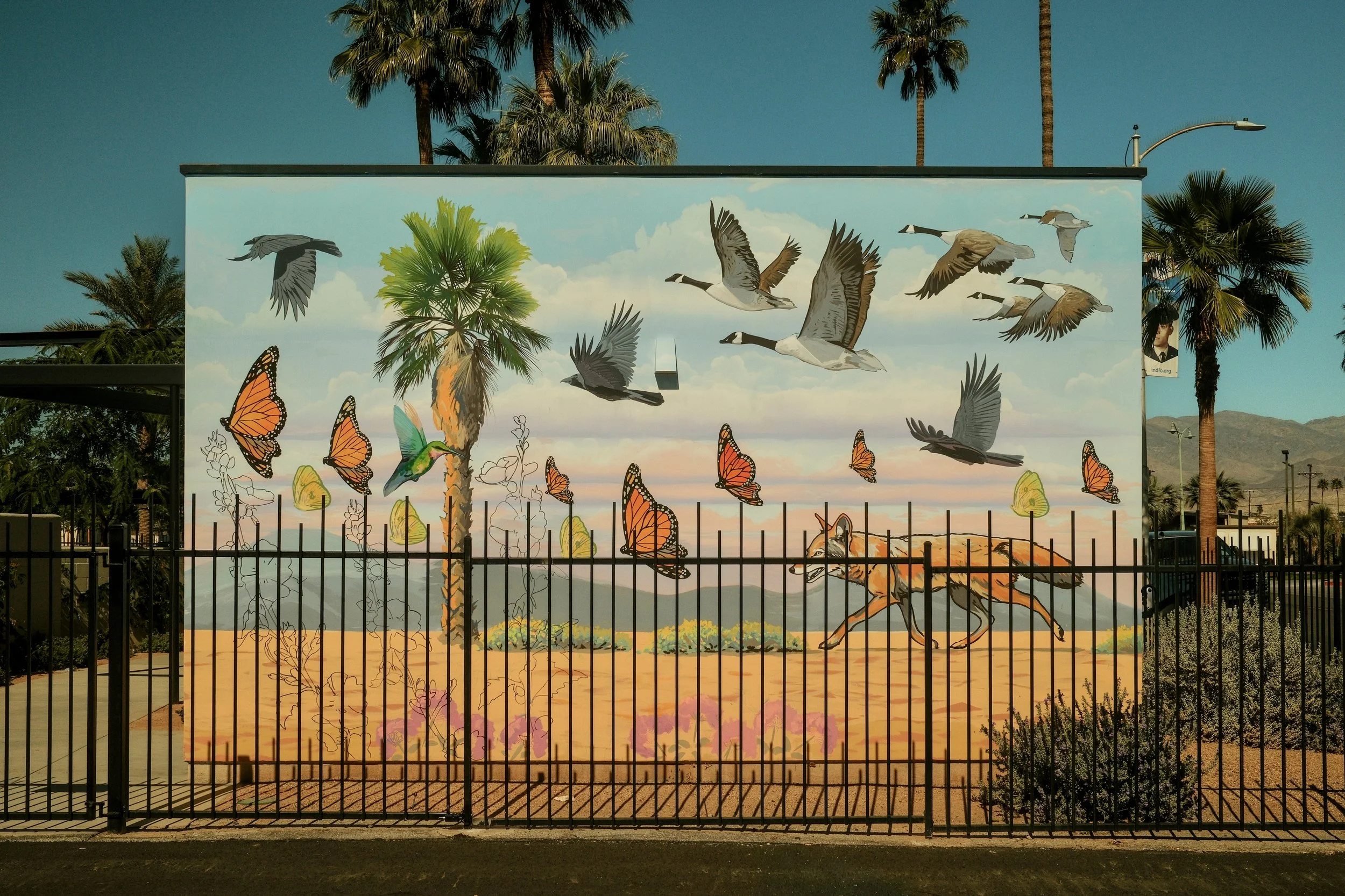 A colorful mural depicting various birds, butterflies, a coyote running on the ground, and a tree, with a mountain landscape in the background.
