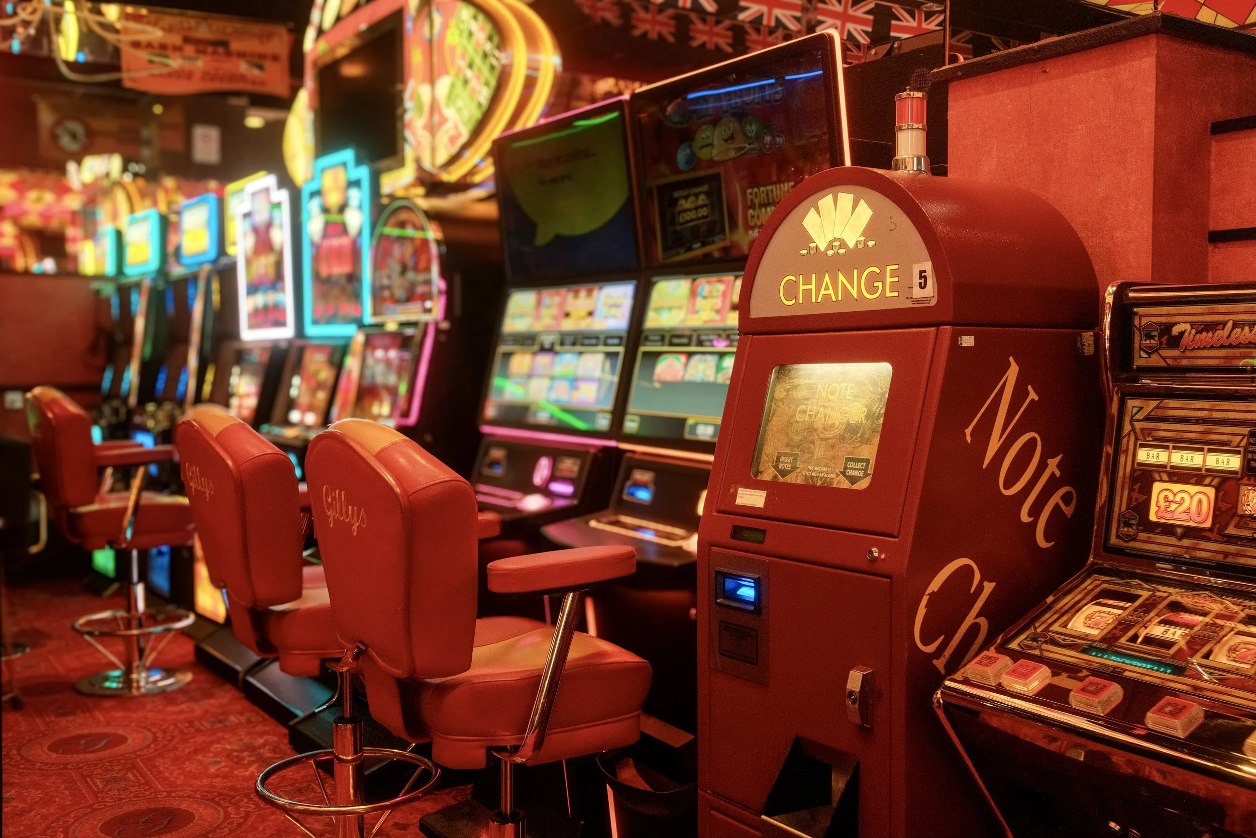 Brightly lit row of slot machines with red bar stools in front, in a casino with colorful neon lights and flags hanging overhead.