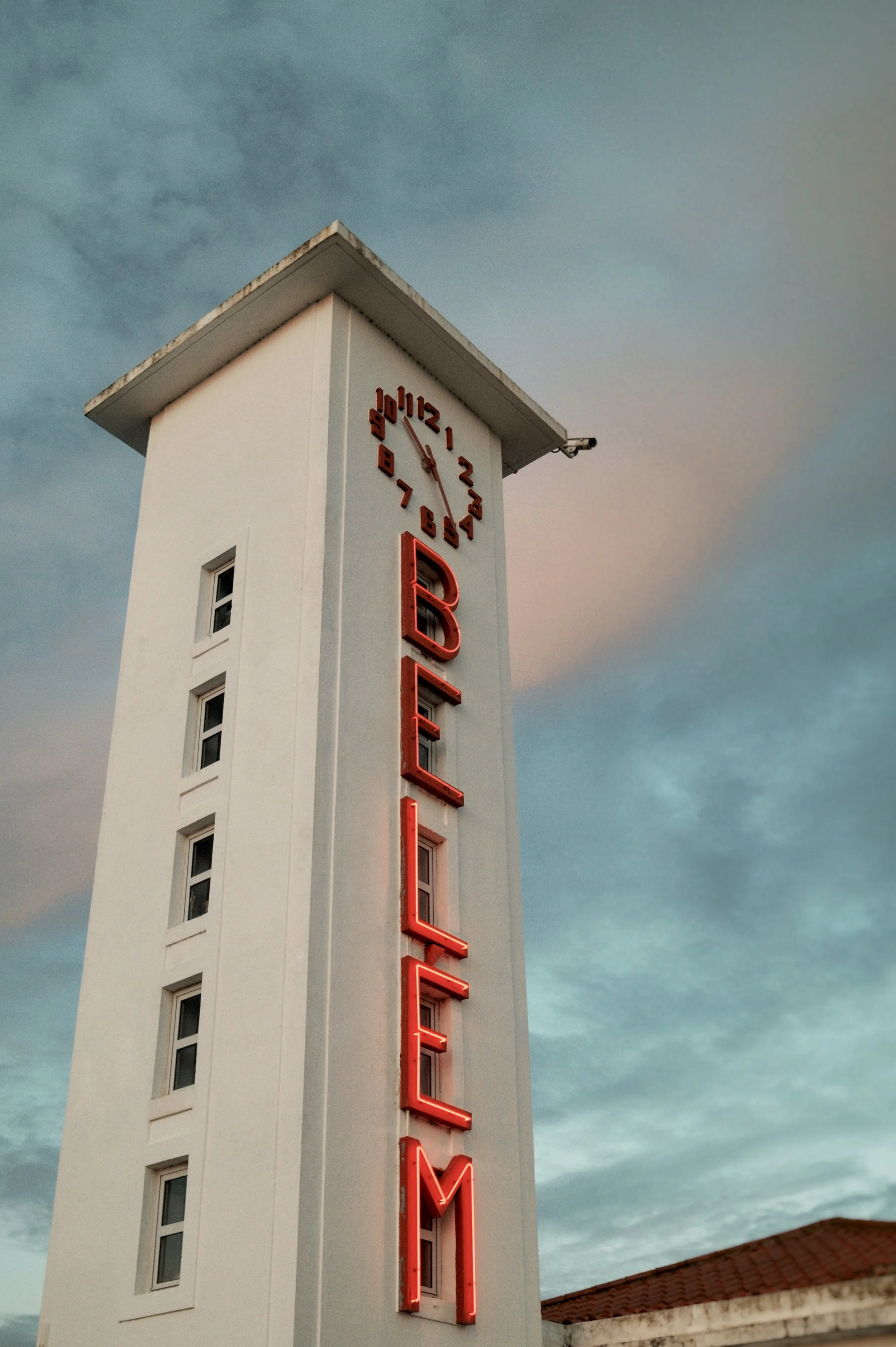 A tall white hotel sign tower with a neon sign reading 'HOTEL M' and a clock showing approximately 5:50, set against a cloudy sky during dusk.