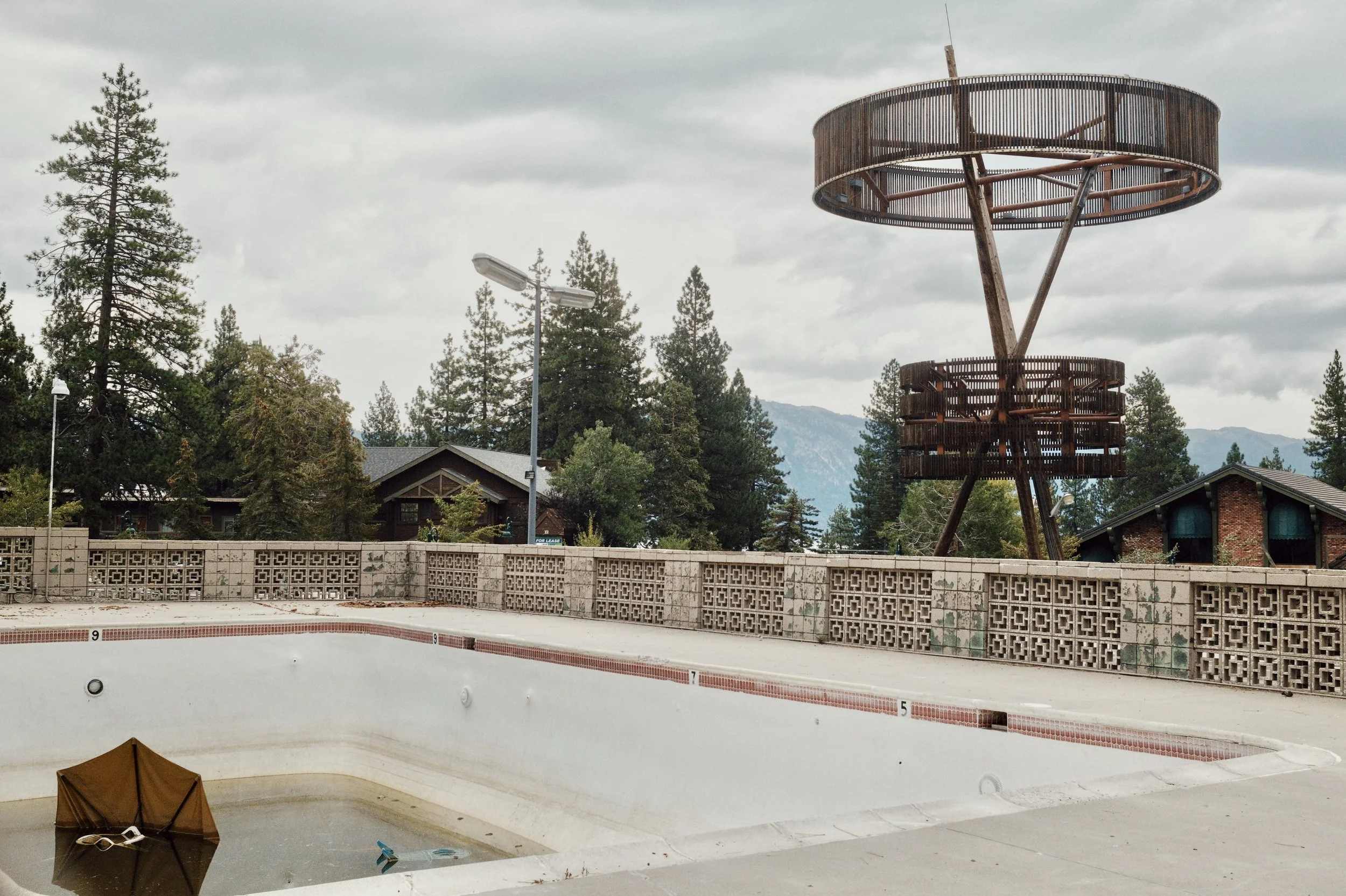 An empty, abandoned swimming pool with a brown umbrella floating upright in the water, surrounded by a concrete deck and a decorative brick wall. In the background, there are trees, a few houses, L-shaped street lamps, and a large rusted metal struct
