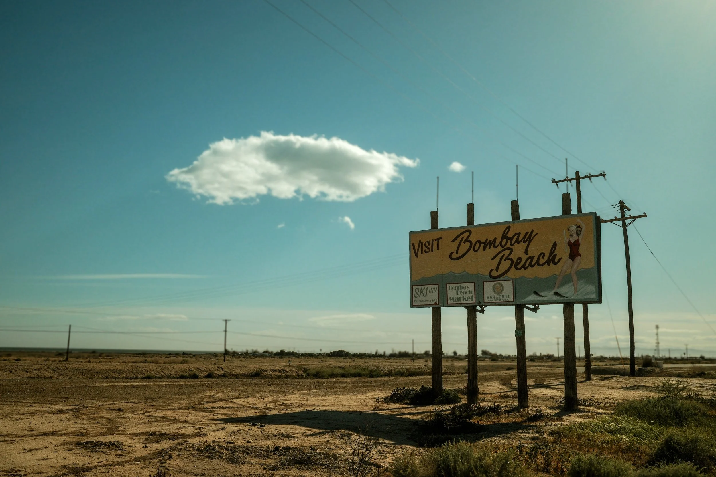 A roadside billboard advertising Bombay Beach with logos for ski inn, Bombay Beach Market, and bar & grill, set in a desert landscape under a blue sky with a single cloud.