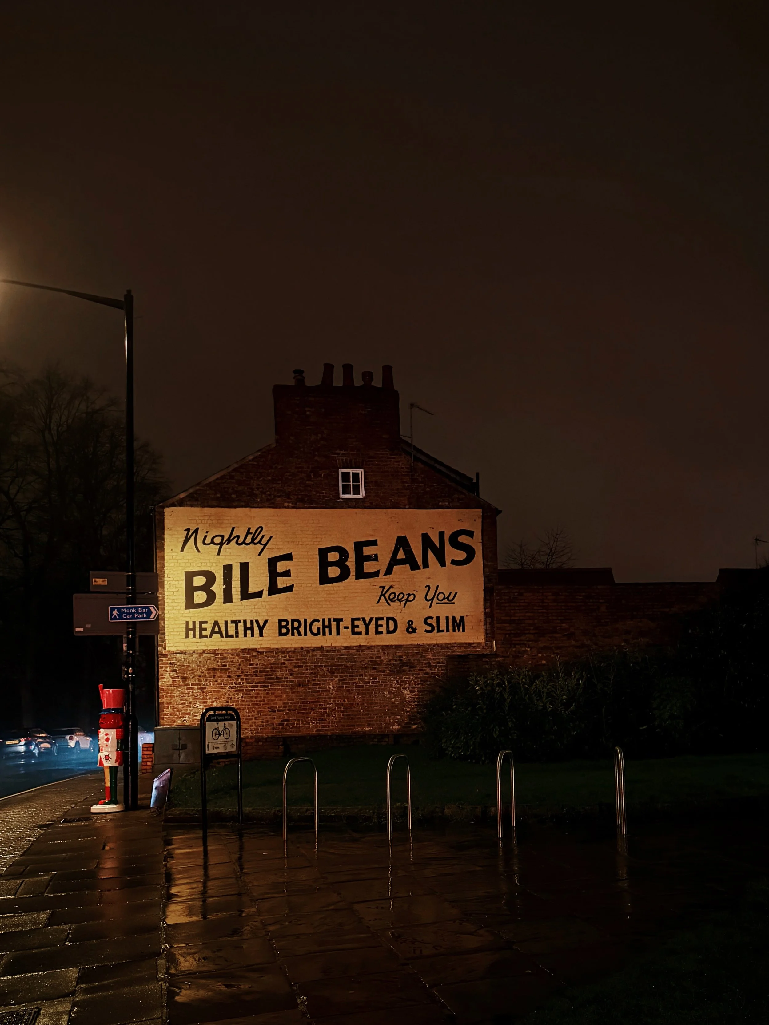 A vintage brick building with a large sign advertising Bile Beans, a medicinal product, illuminated by streetlights at night. The sign promotes Bile Beans as keeping you healthy, bright-eyed, and slim.