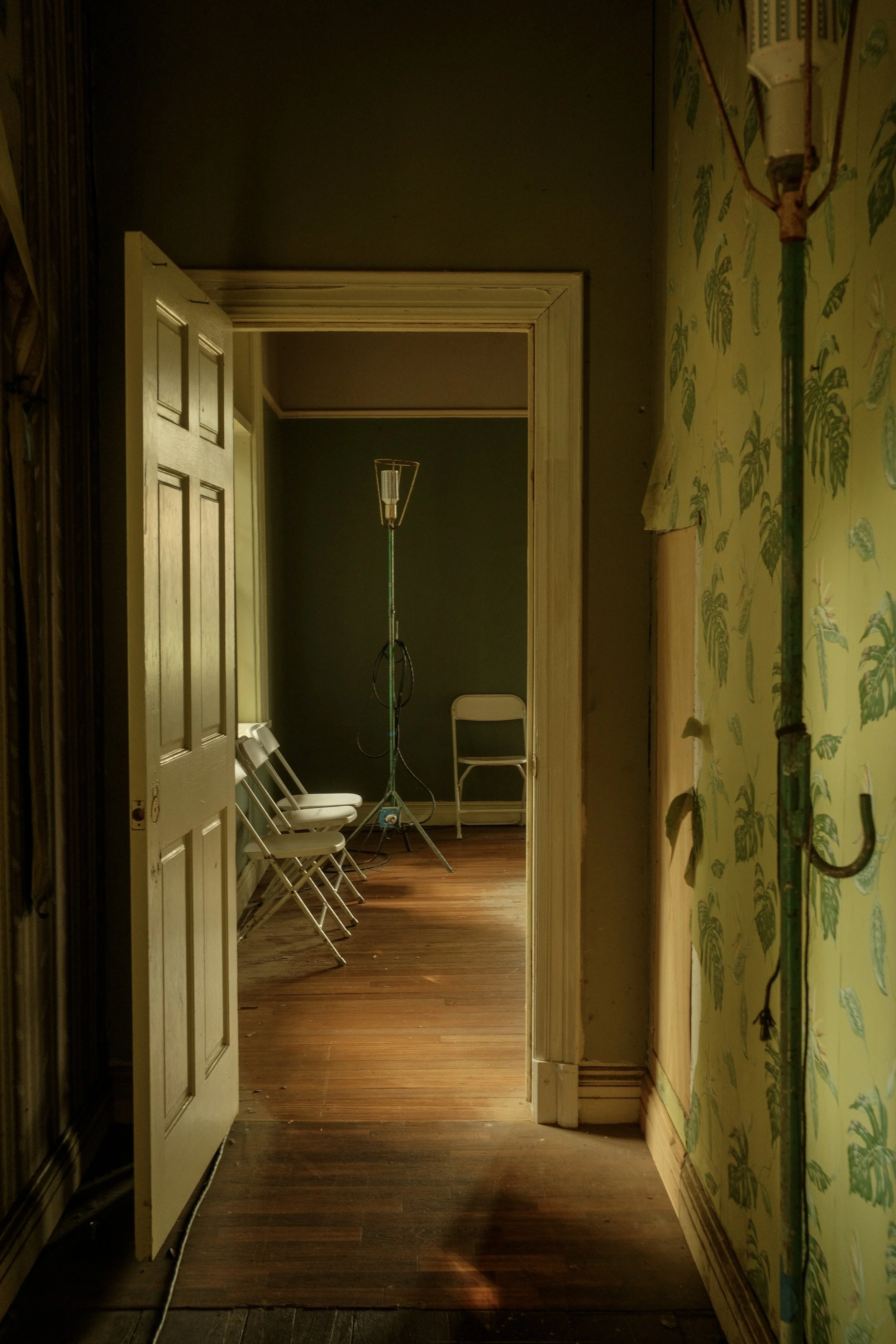 An interior room with wooden flooring, yellow patterned wallpaper on the wall, and a glass IV drip stand in the center. Four white folding chairs are arranged along the left wall, with one chair positioned separately in the back corner. A closed whit