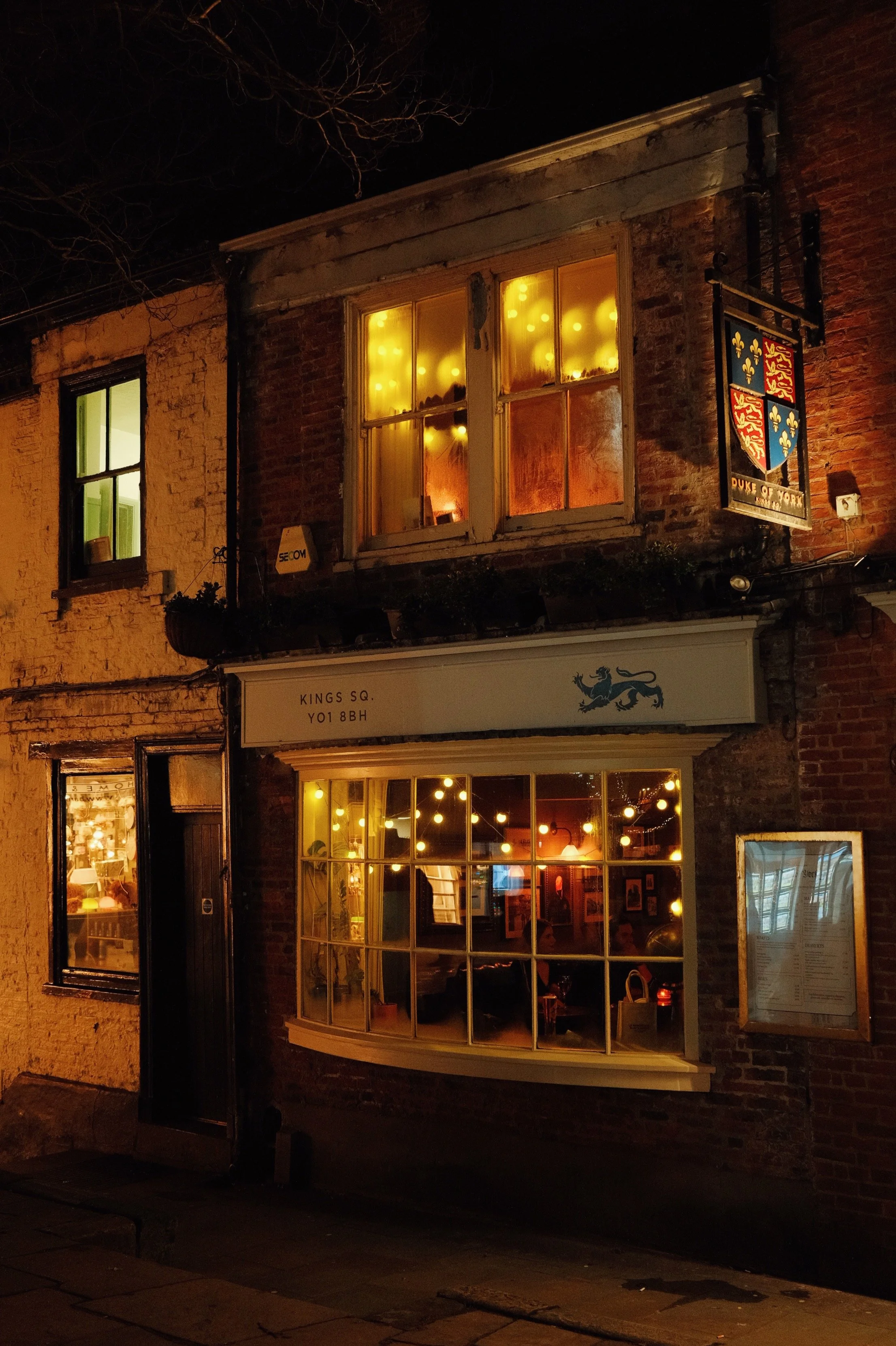 Nighttime exterior view of a brick building with illuminated windows, displaying warm yellow and orange lights, and a sign for a restaurant or pub. The building features a shield-shaped crest with a lion and floral patterns.
