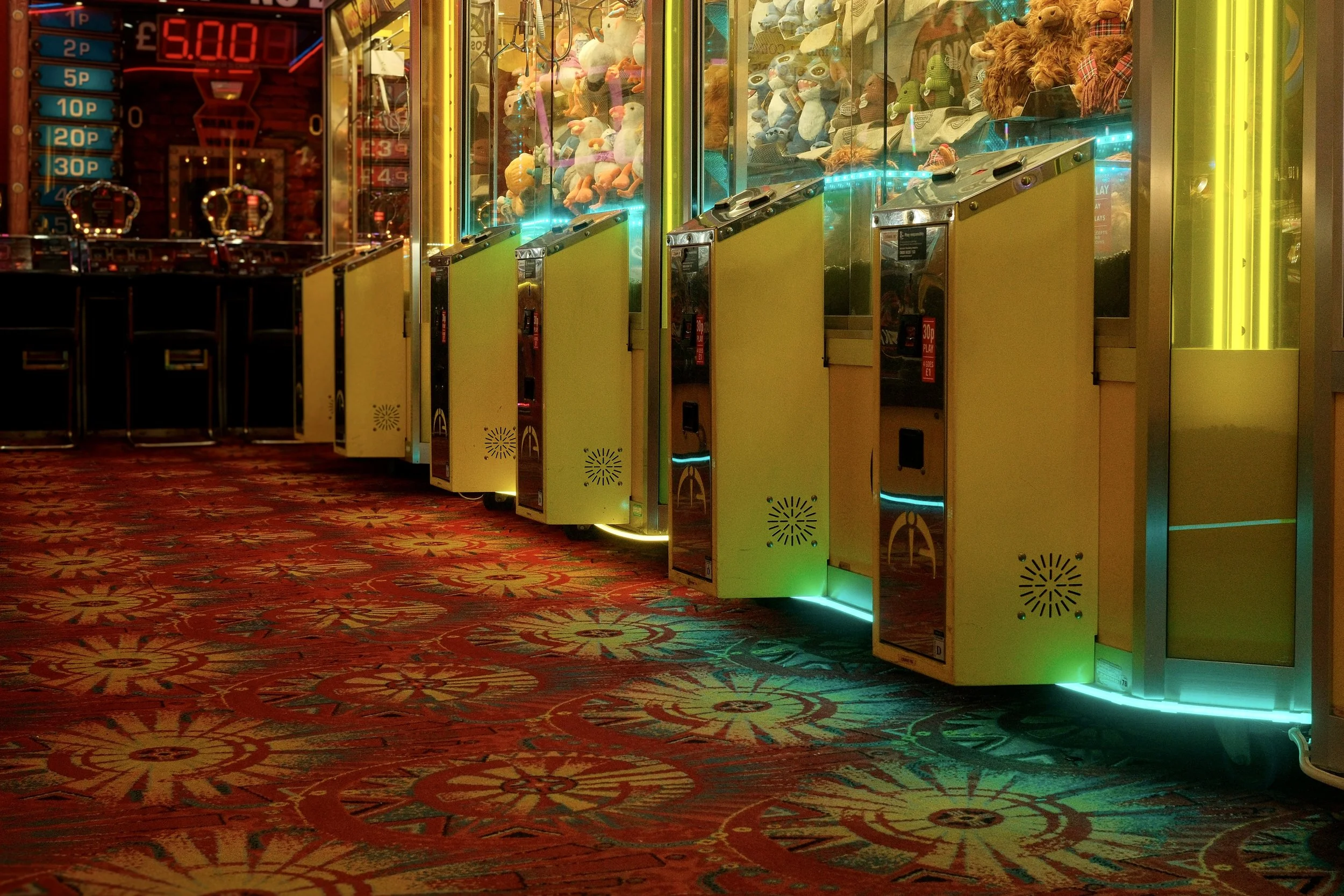An arcade with claw machines filled with plush toys and a gaming machine with a digital payout display, set on a patterned carpet in a dimly lit gaming area.