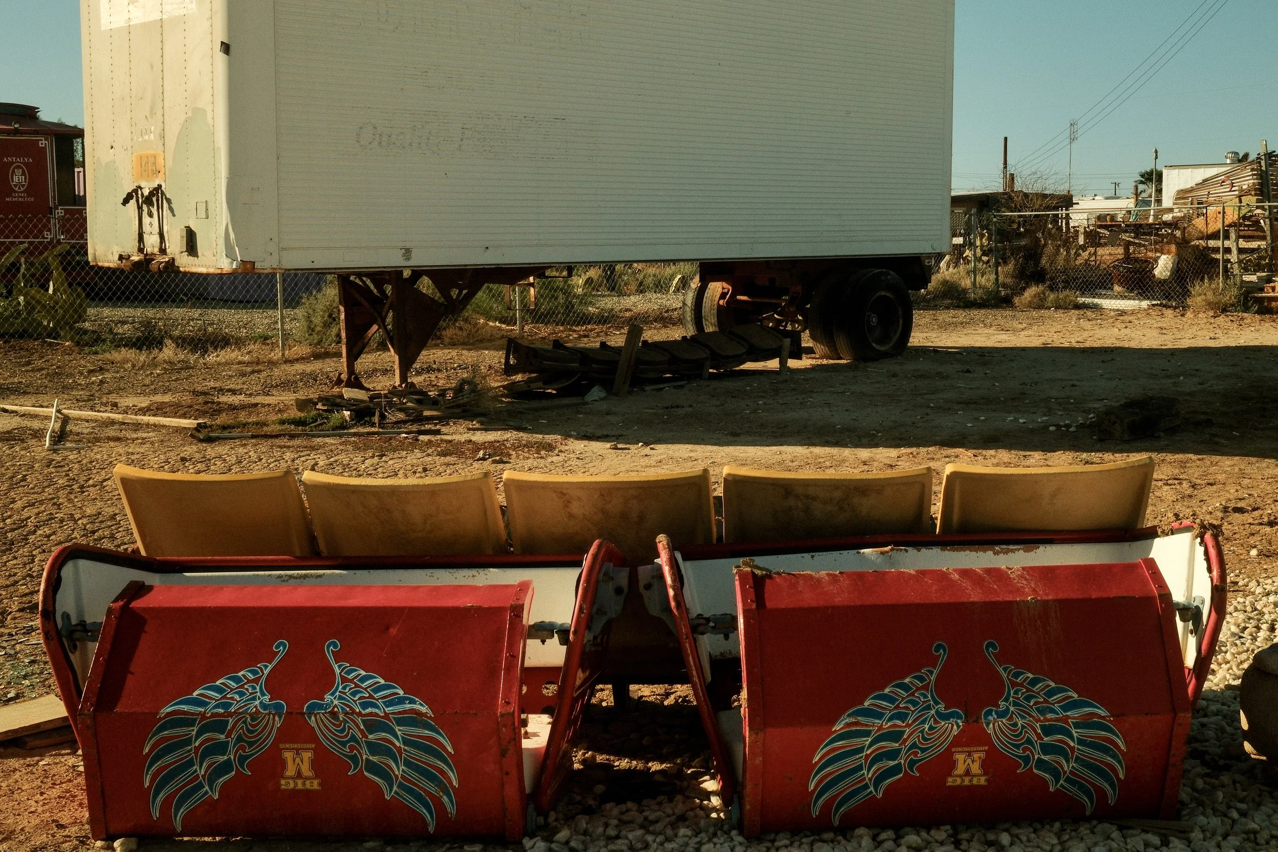Old bumper cars with winged insect graphic, yellow seats in front of a mobile trailer on dirt ground, with miscellaneous junk and a chain-link fence in the background.