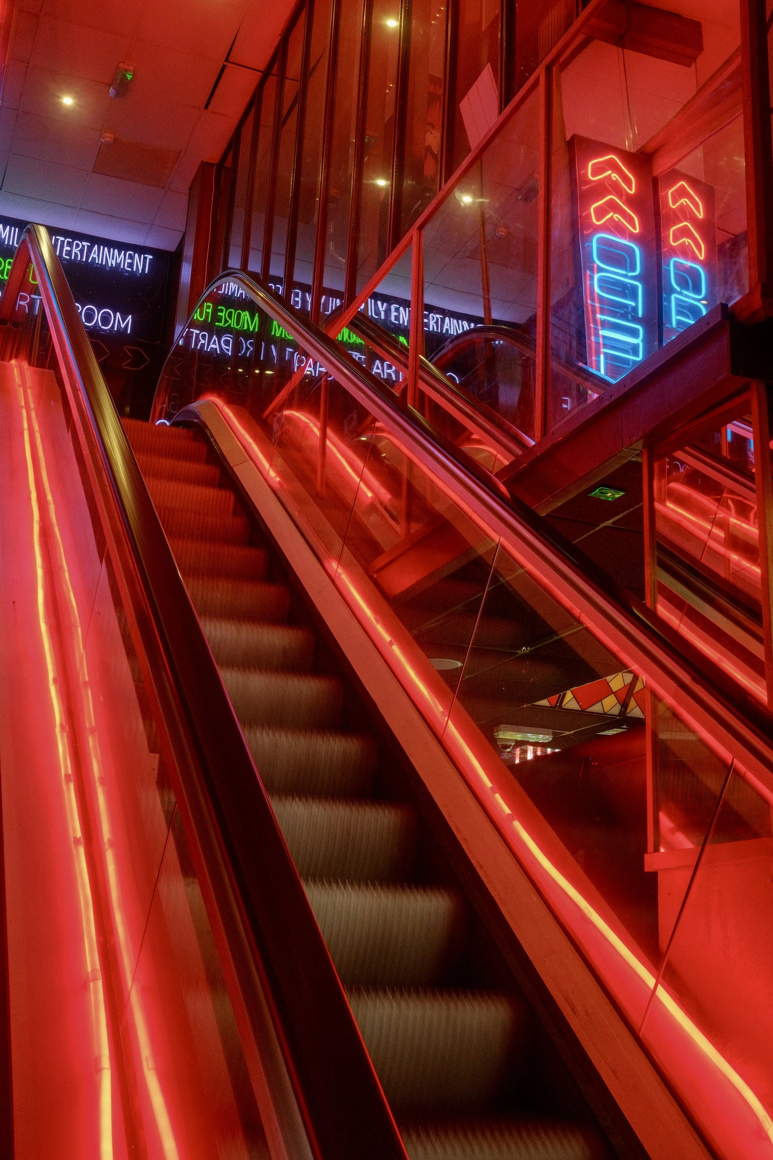 Neon signs in a building with red and blue lights, showing arrows pointing up and the word 'ENTRANCE,' and other illuminated signage reflected in glass panels.
