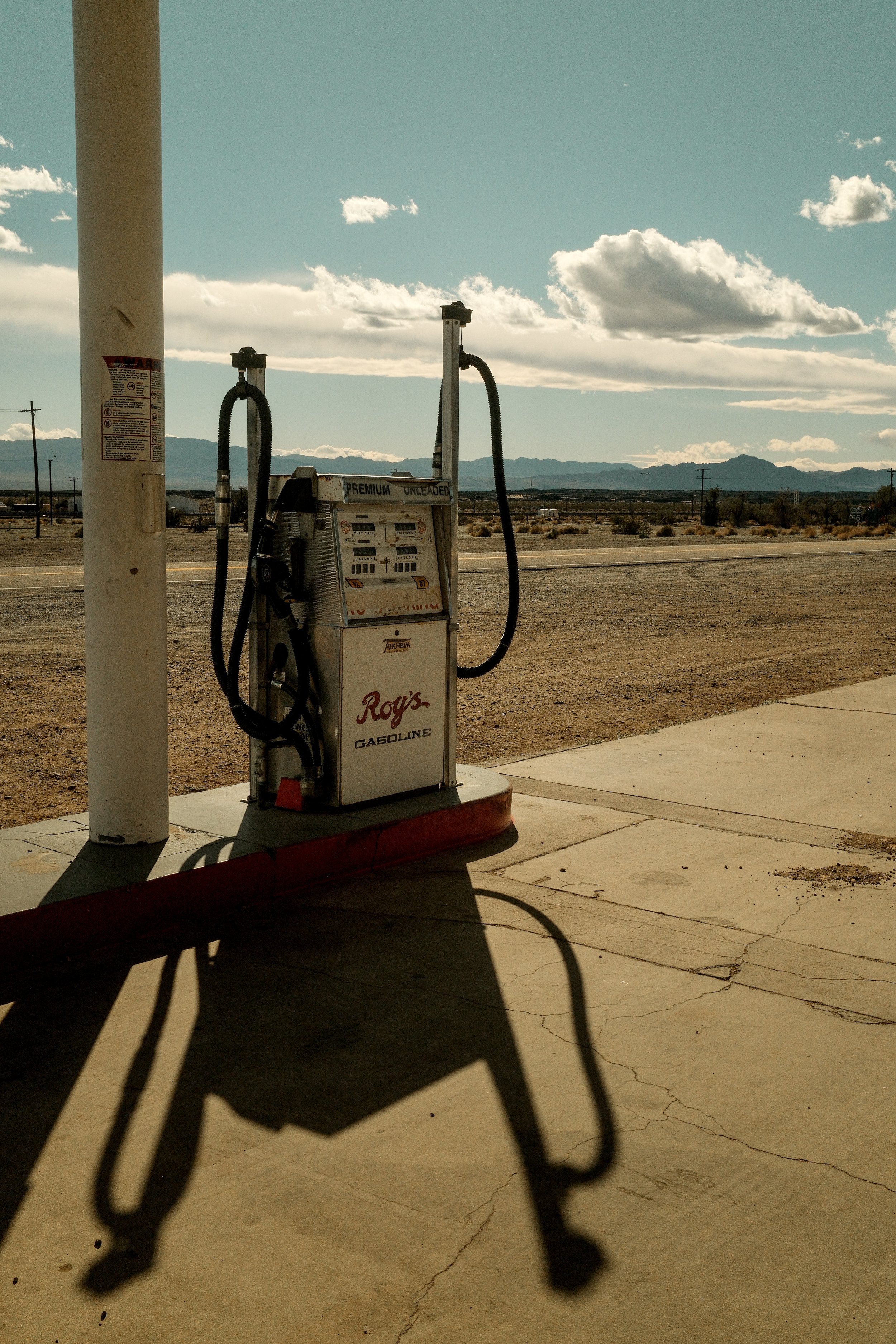A gas pump on a sidewalk outside, with a desert landscape and mountains in the background under a partly cloudy sky.