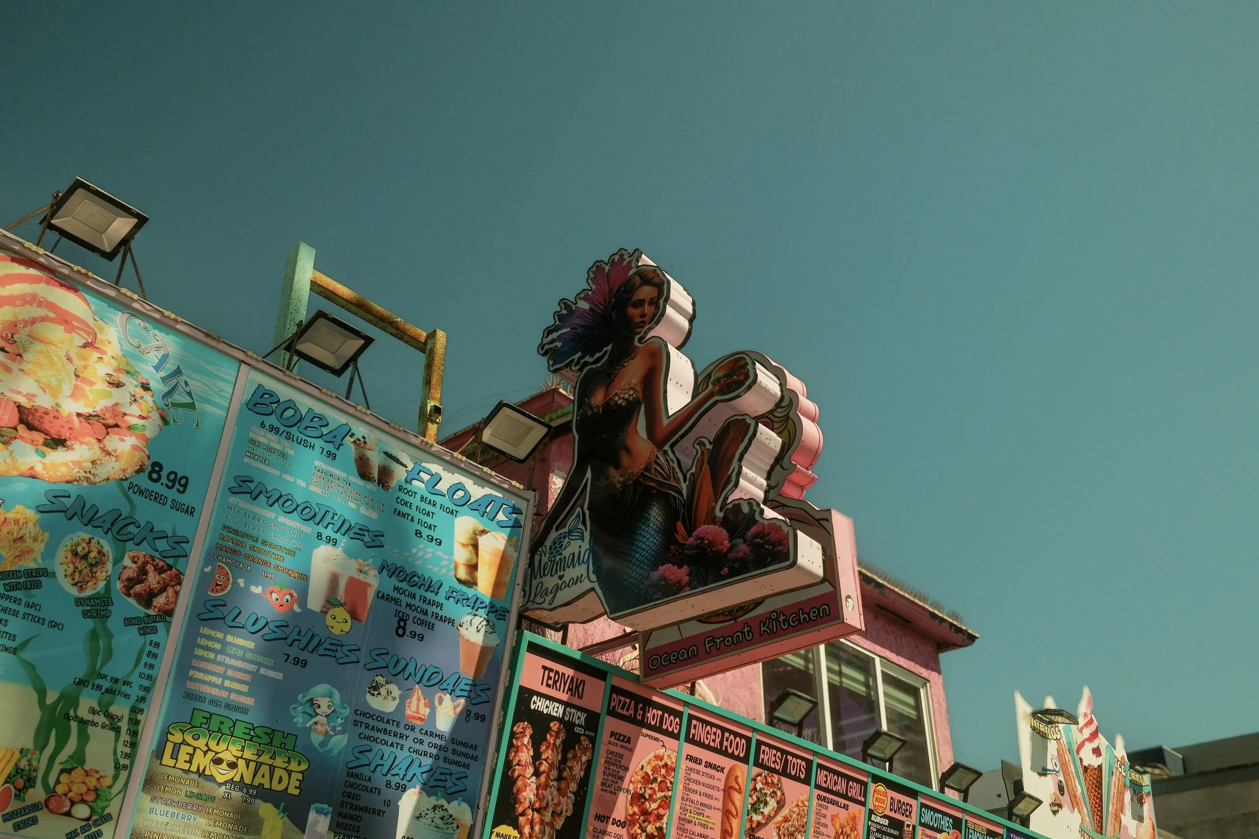 Colorful food stand with a large illustrated mermaid sign and menu boards listing various fried foods, smoothies, and desserts against a clear sky.
