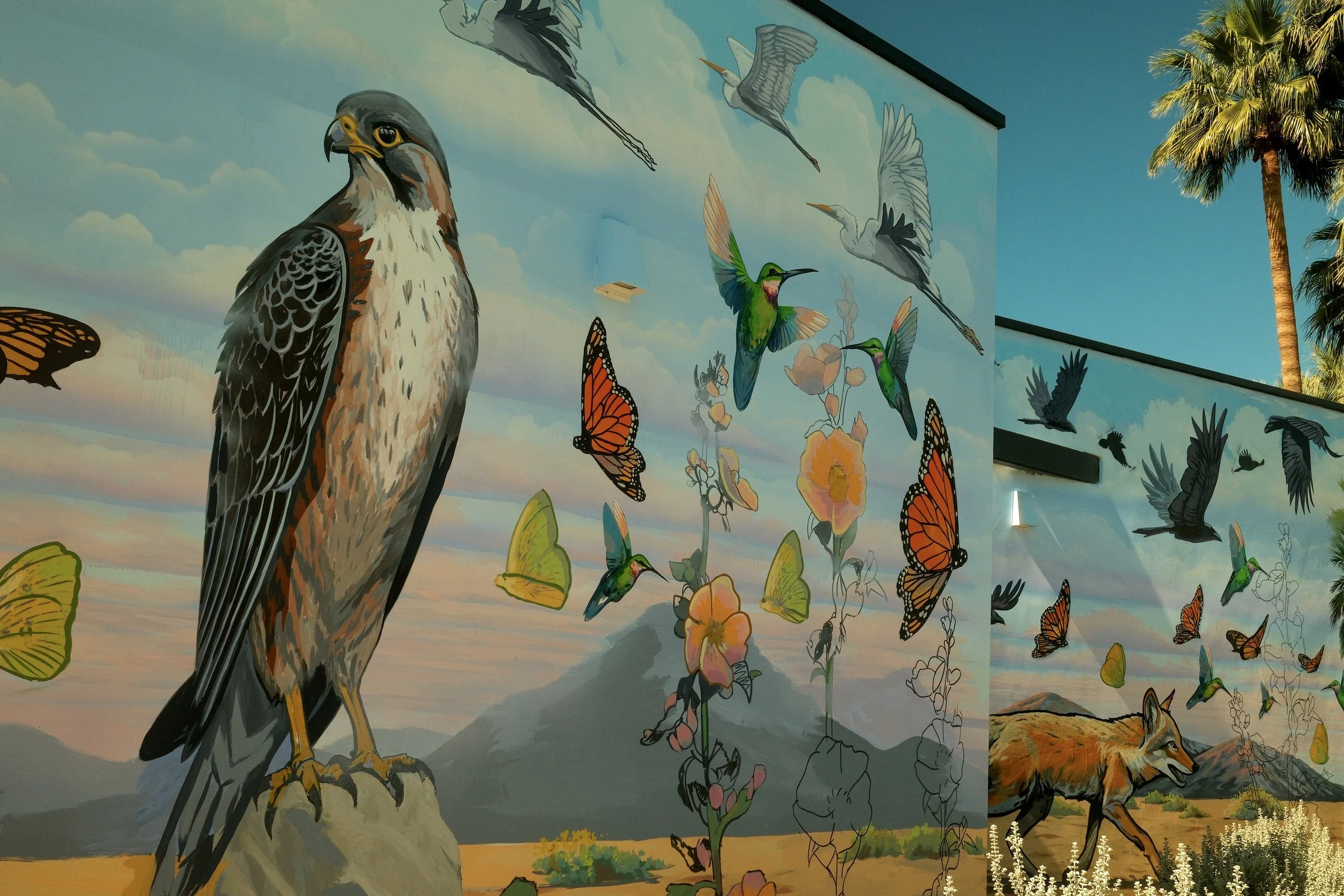 A mural depicting a hawk perched on a rock, surrounded by flying hummingbirds, butterflies, and flowers, with mountains and a cloudy sky in the background.