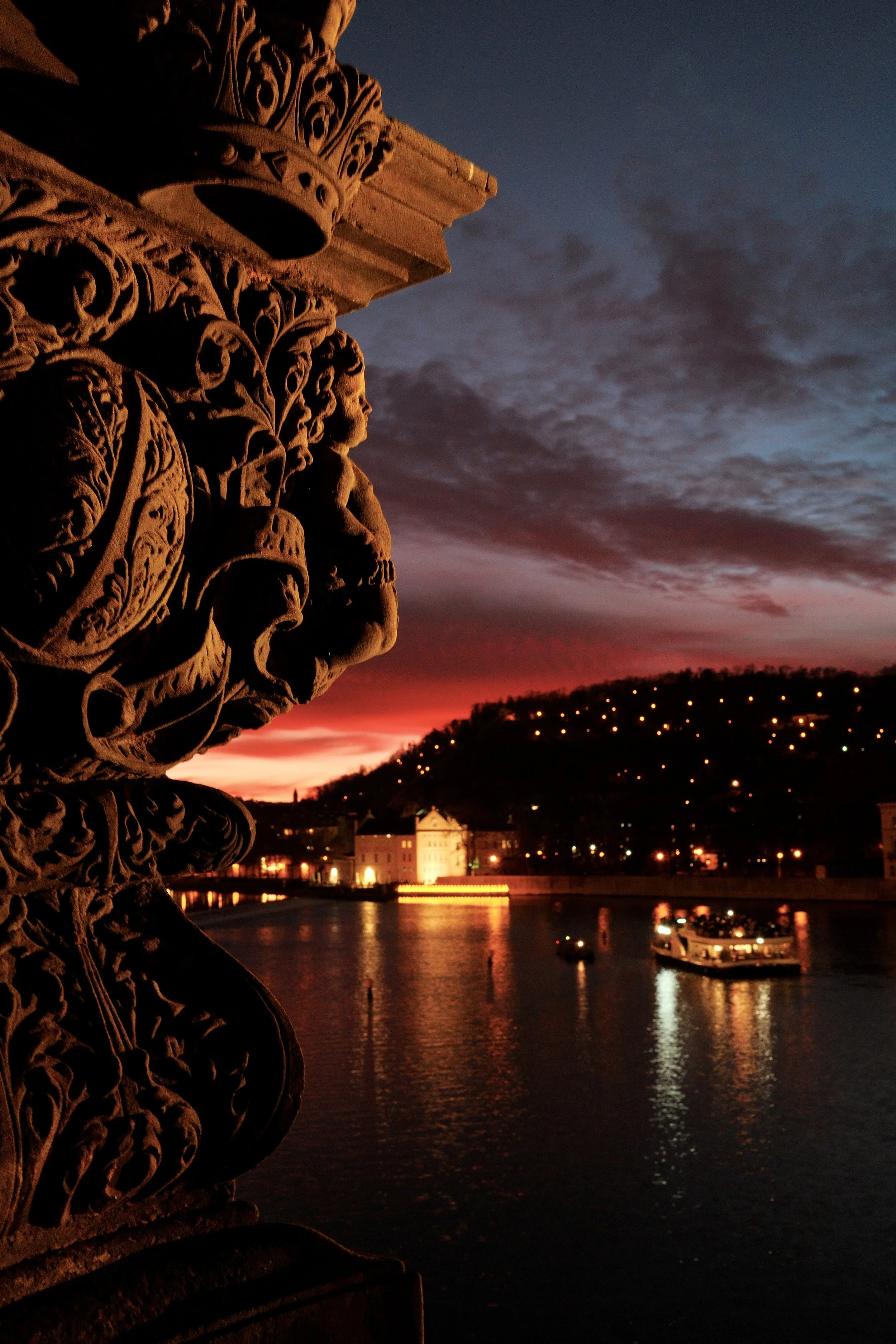 A close-up of ornate stone carving on a bridge railing at sunset, with a river, boats, and a hill with lights in the background.