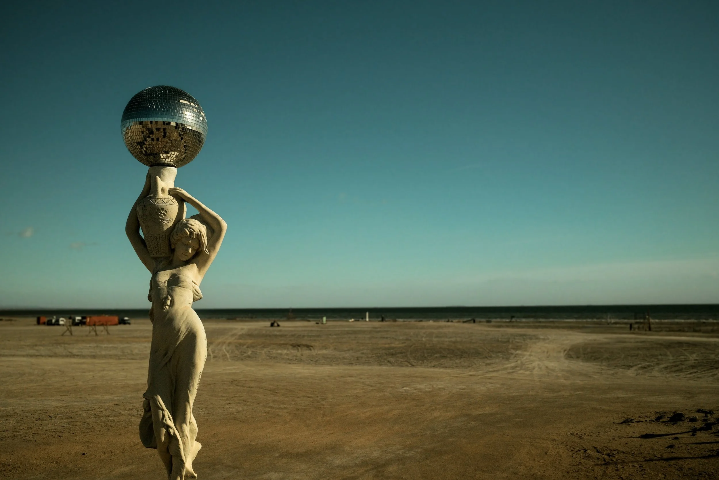 A sculpture of a woman holding a large vase on her head with a shiny disco ball orb balanced above it, set against a wide, empty beach under a blue sky.