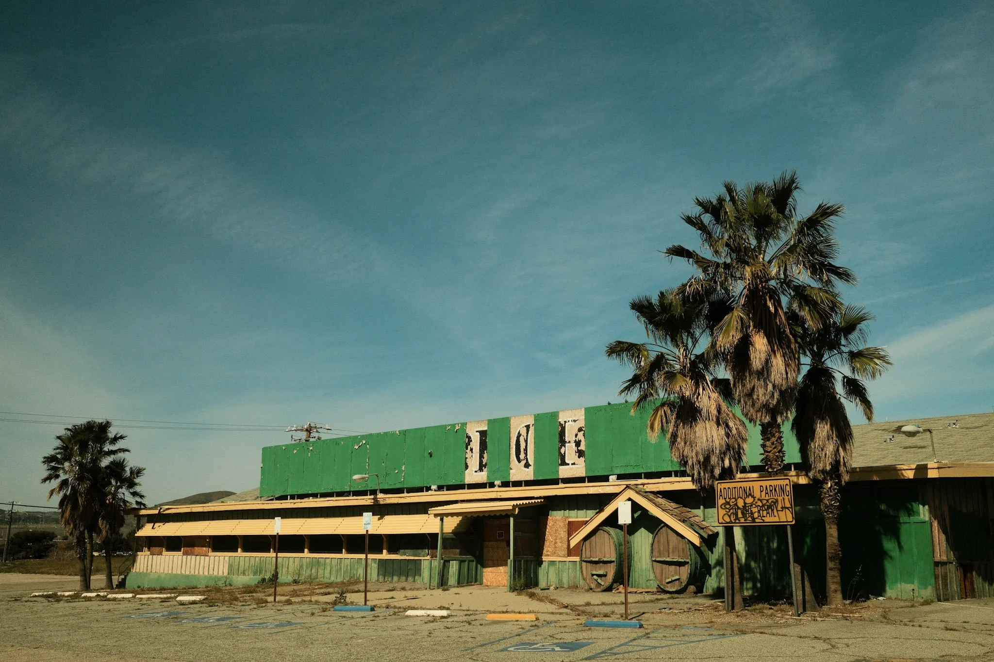 Abandoned restaurant building with green faded siding and a large sign, two palm trees, empty parking lot with designated handicapped spots, and a yellow parking sign that says 'Additional Parking'. The sky is clear with a few clouds.