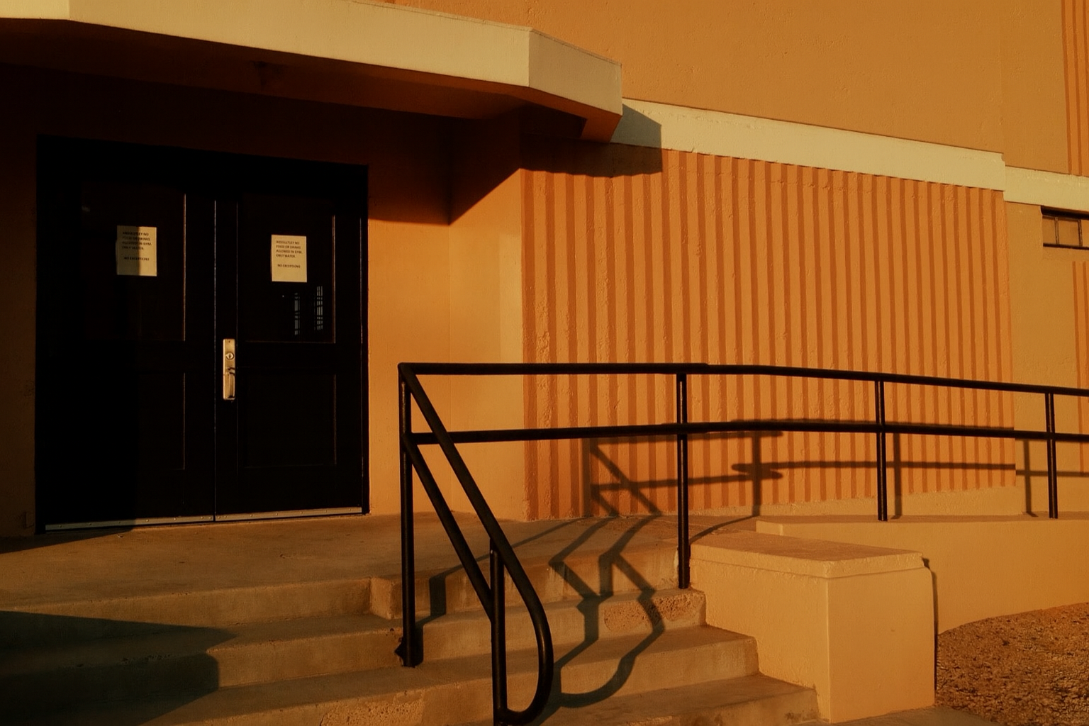 Dark entrance door with signs, handrails on stairs leading up to the door, and an textured wall on the right side, illuminated by warm lighting.