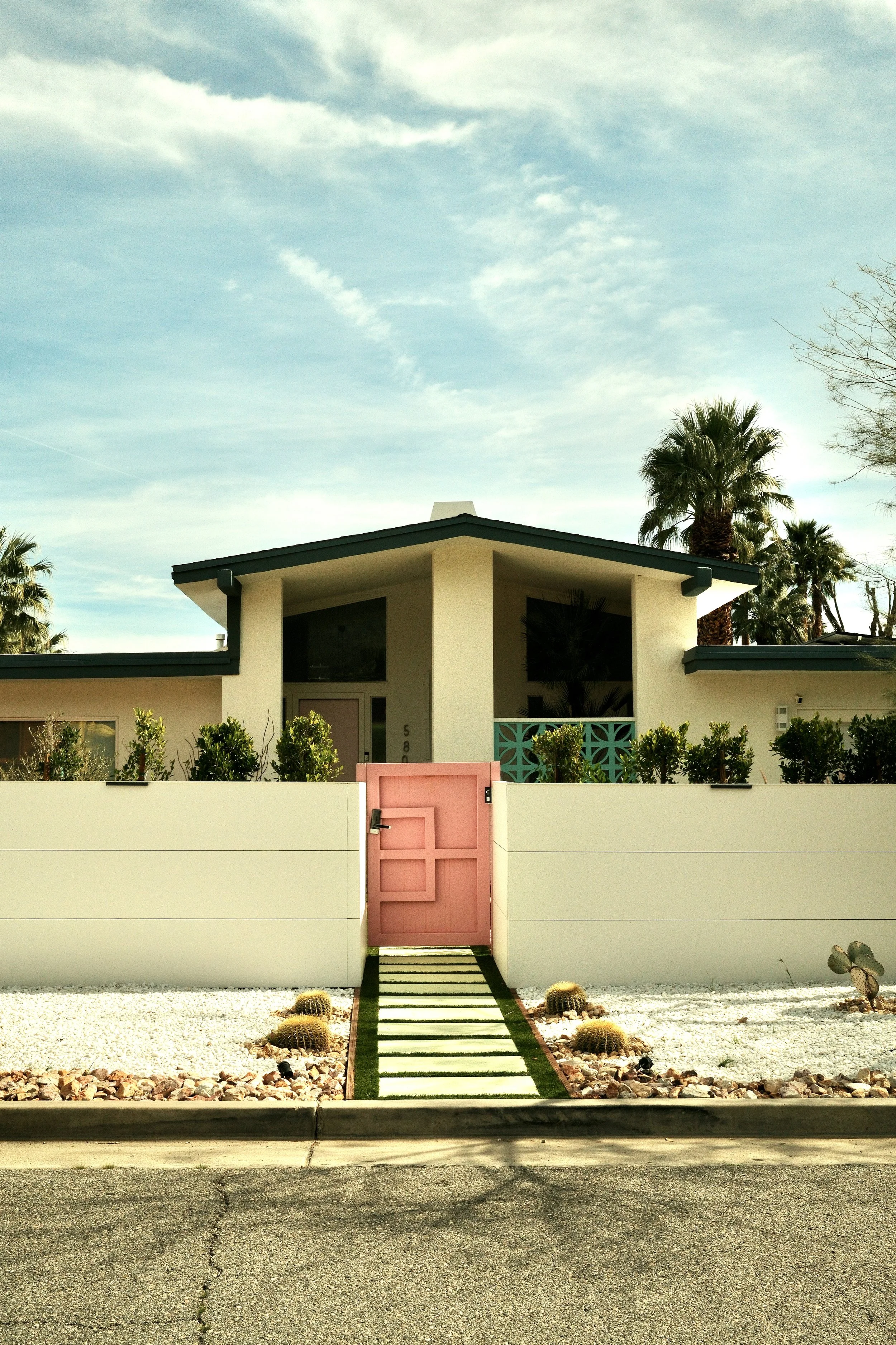 Modern house with white walls, a pink gate, and desert landscaping, including cacti and stone gravel, under a partly cloudy sky.