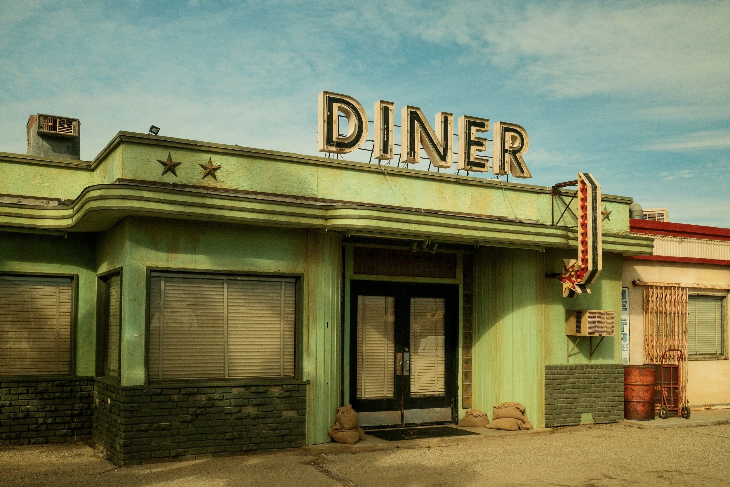 Old, weathered diner building with a large illuminated 'DINER' sign on top and an arrow-shaped neon sign on the side, featuring closed shutters and a green facade.