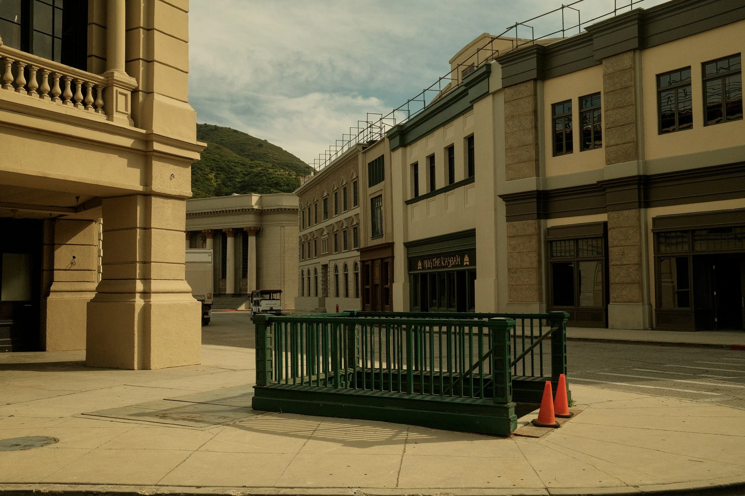 Empty city street with multi-story buildings, a green barricade with orange cones, and a green mountain in the background.