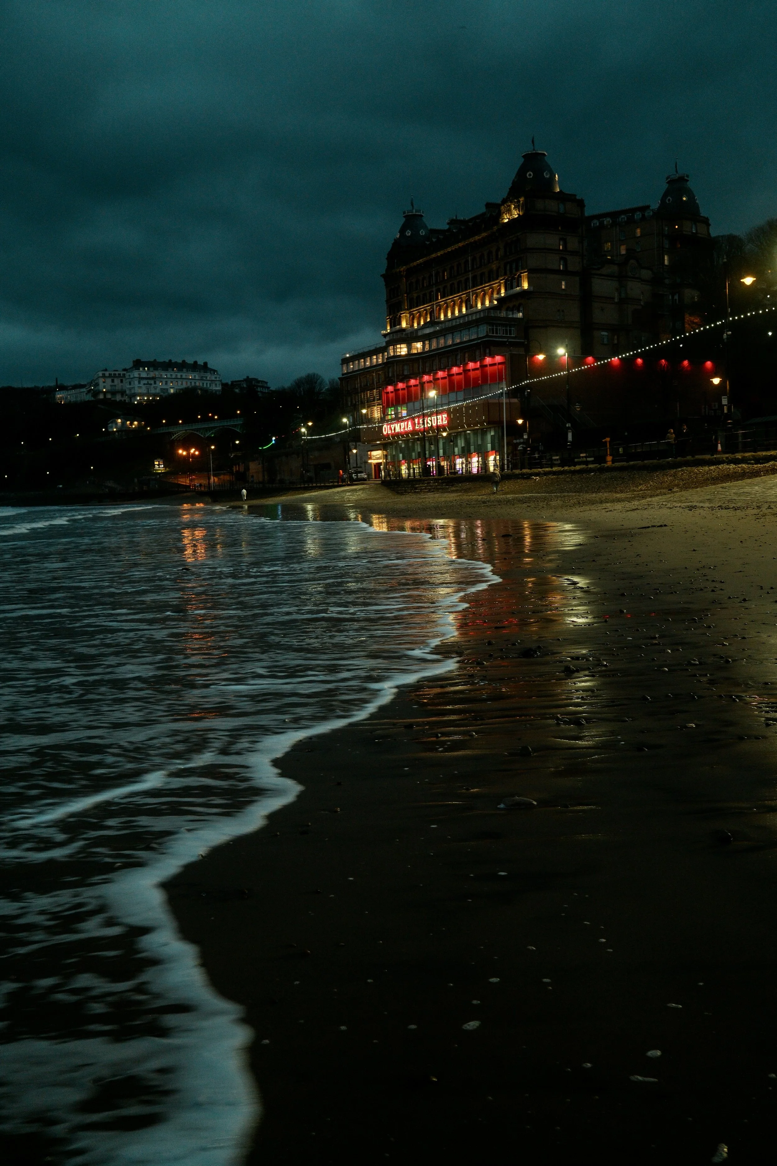 A nighttime view of a beach with calm water and gentle waves, a large ornate casino building with lights on, and a dark cloudy sky overhead.